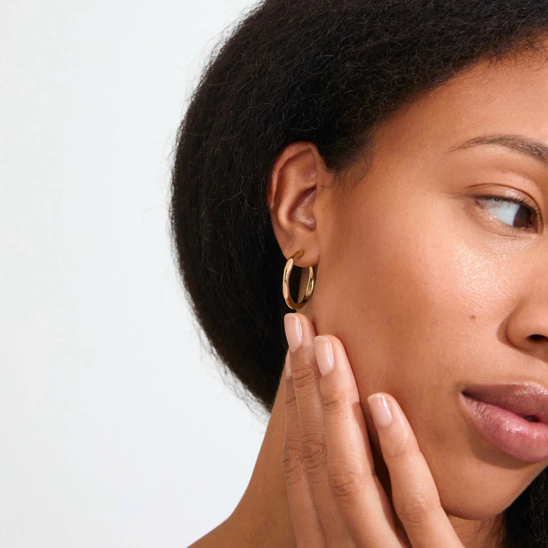 A close-up of a woman wearing a thin gold hoop earring, with her hand resting against her cheek. The background is a clean, solid off-white studio wall.
