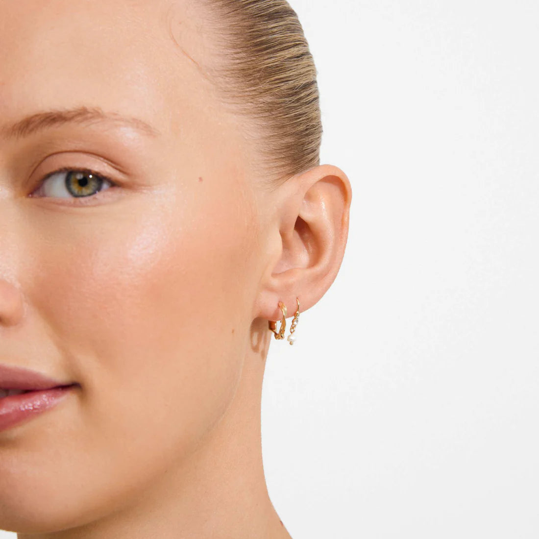 Close-up of a woman's ear wearing a pair of small gold hoop earrings against a clean white background.