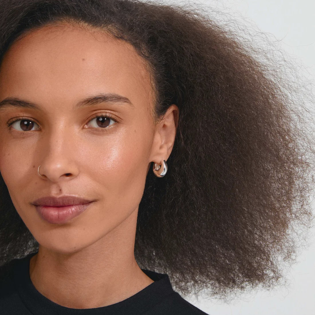 A close-up portrait of a woman with dark curly hair wearing a small silver crescent moon hoop earring, set against a plain light gray background.