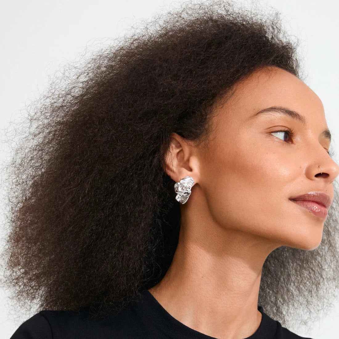 A close-up portrait of a woman with voluminous dark curly hair wearing a silver, diamond-like textured earring and a black top against a light gray background.