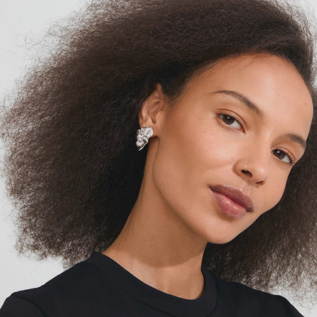 A close-up portrait of a woman with voluminous dark curly hair wearing a silver, diamond-like textured earring and a black top against a light gray background.
