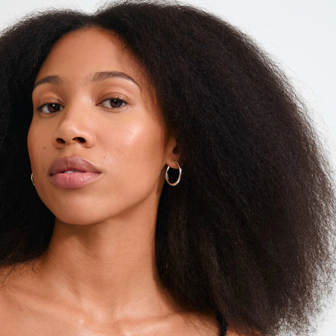 A close-up portrait of a woman with voluminous dark curly hair wearing a delicate gold hoop earring, set against a clean white background.
