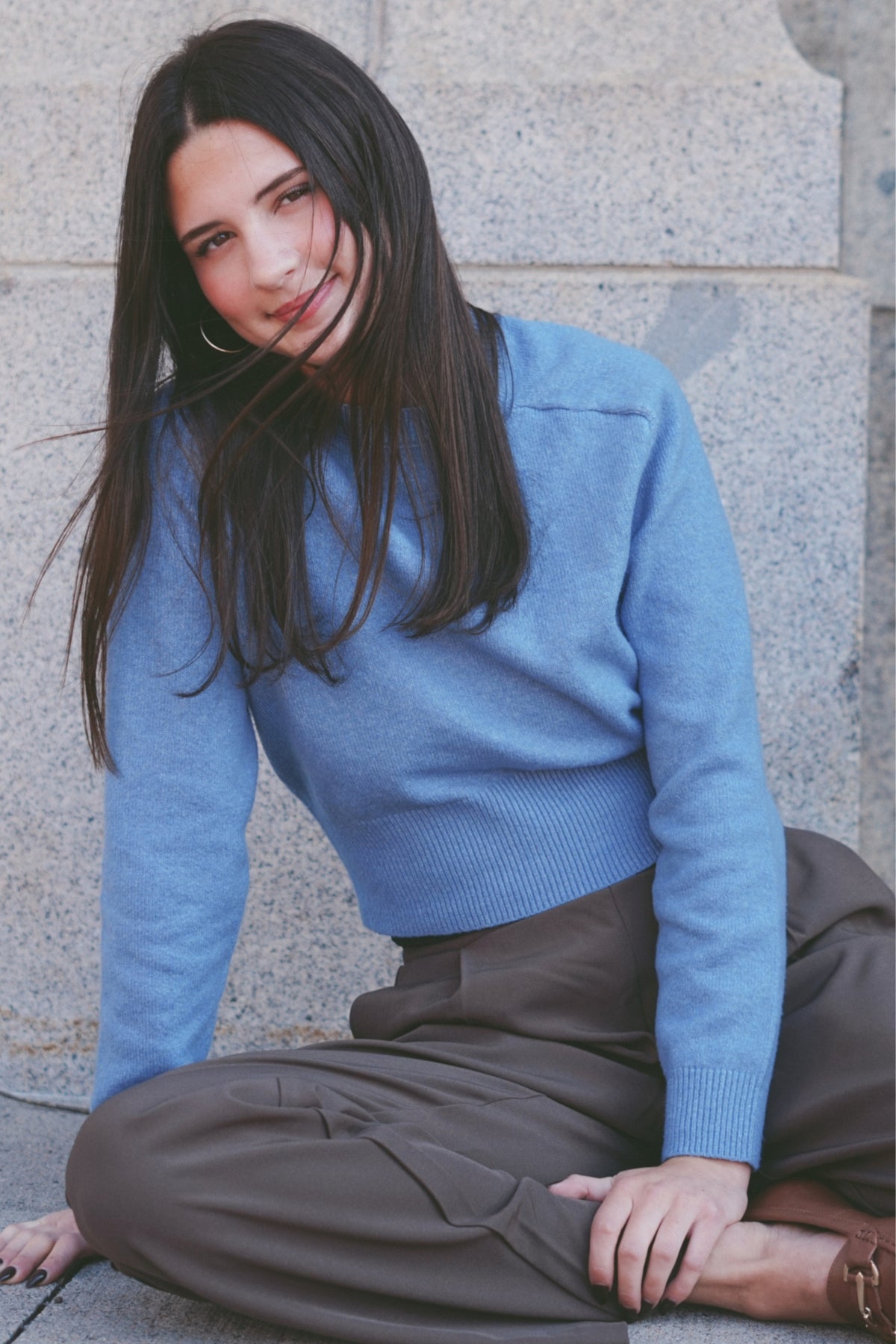 A woman with long dark hair sits cross-legged against a light gray stone wall, wearing a light blue ribbed knit sweater and dark brown pants.