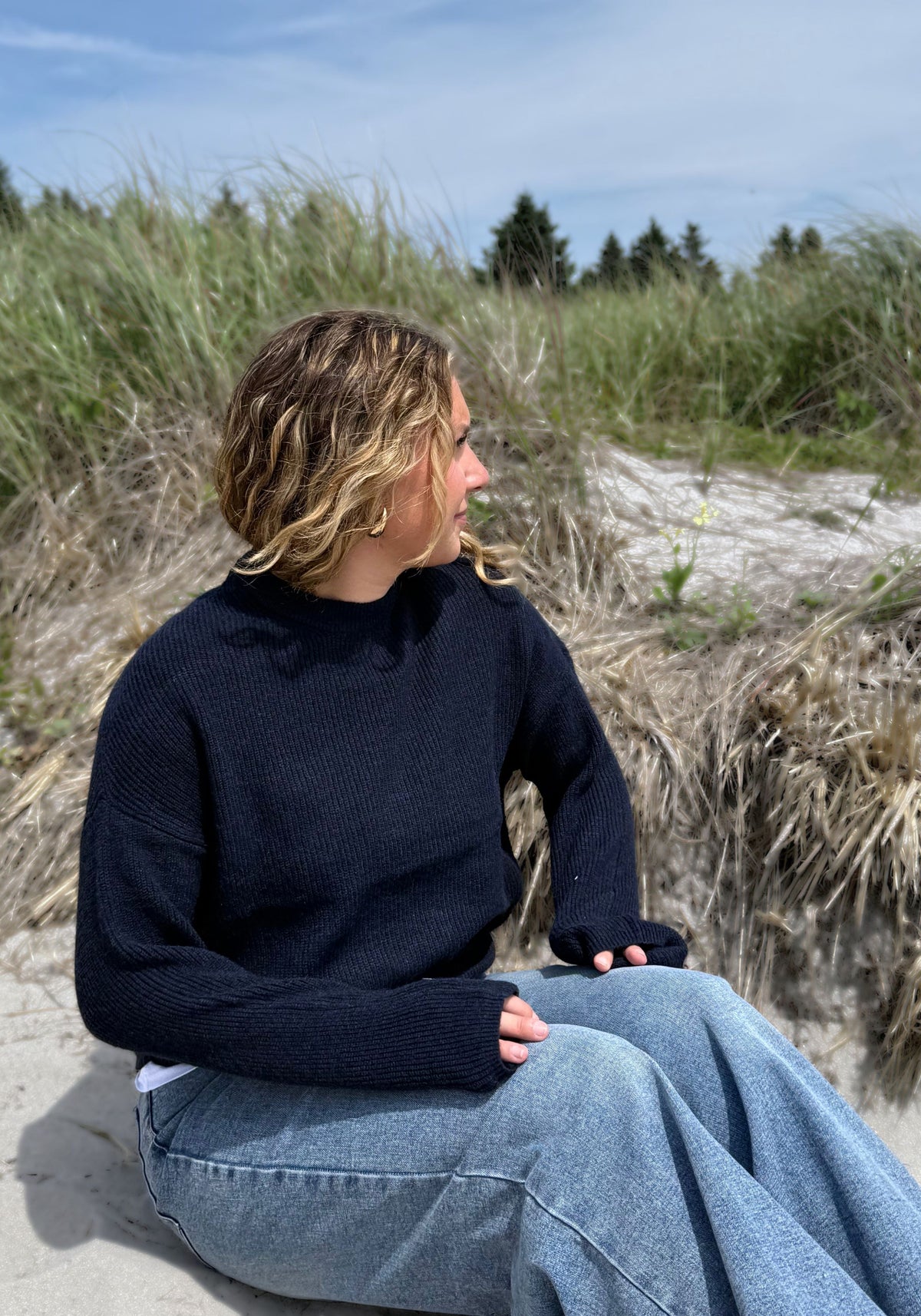 A woman wearing a dark navy ribbed knit mock-neck sweater and light blue wide-leg jeans, with beach grass and trees in the background.