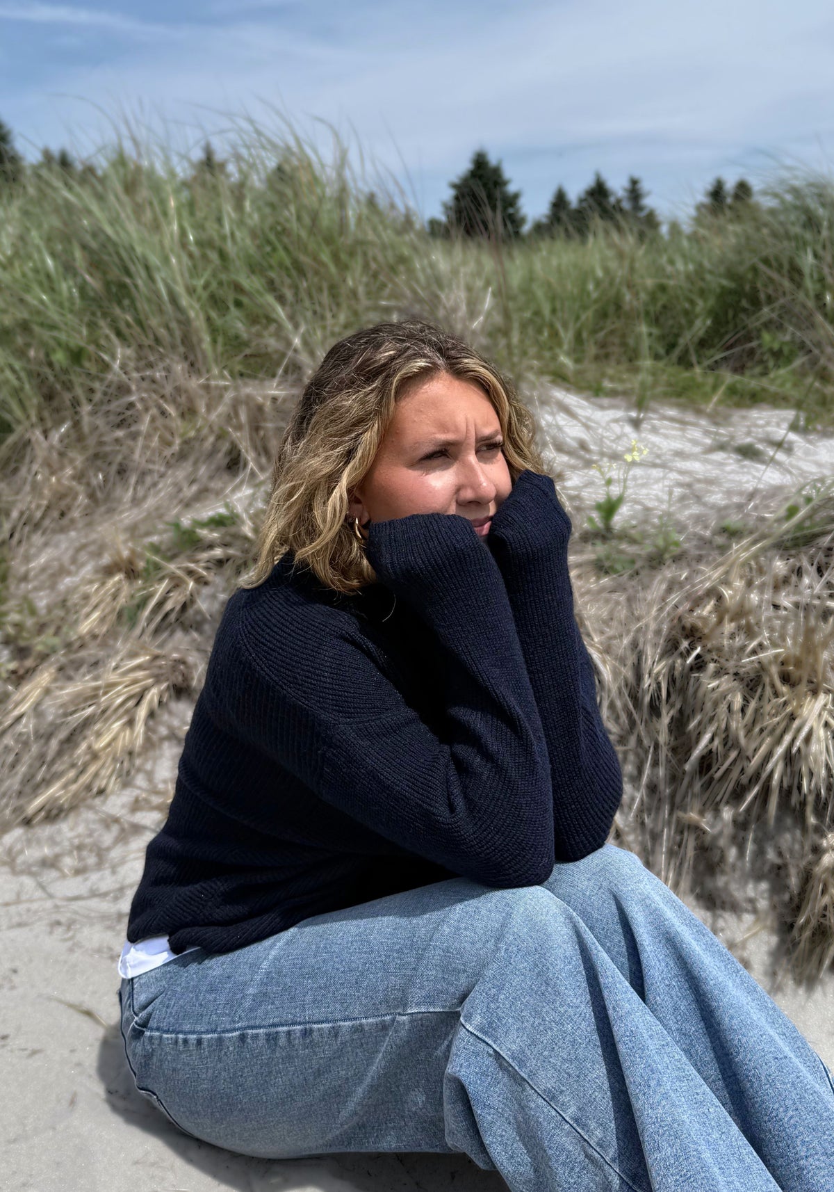A woman sitting on a sandy dune wearing a dark navy knit sweater and light blue wide-leg jeans, with beach grass and trees in the background.