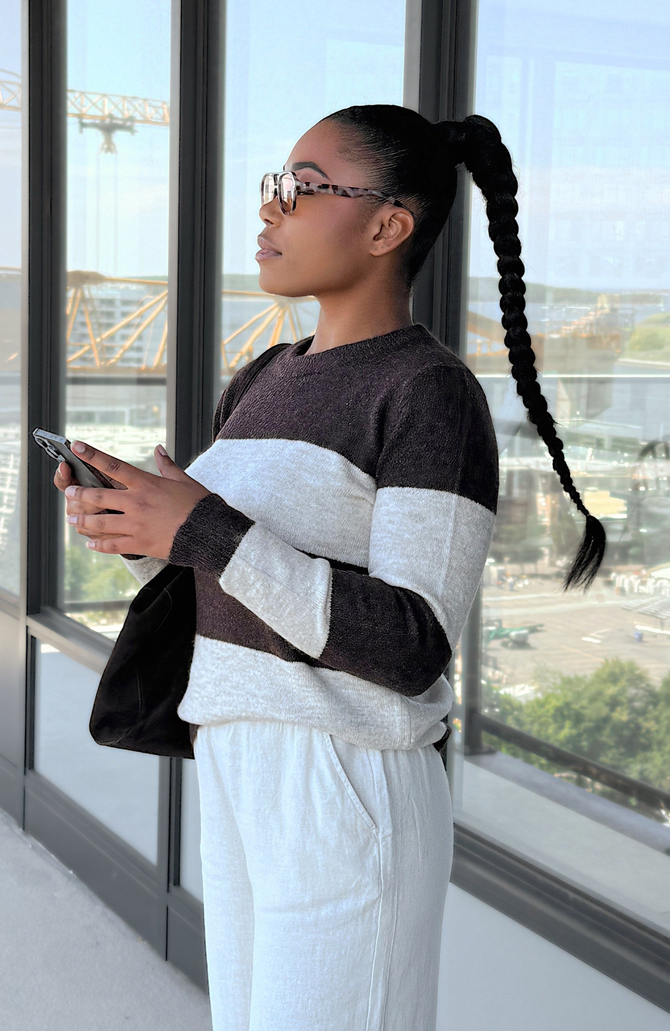 A woman with a long black braided ponytail stands by a large window, wearing a color-block knit sweater with dark brown and cream horizontal stripes, paired with white sweatpants. She is holding a smartphone and carrying a black shoulder bag.
