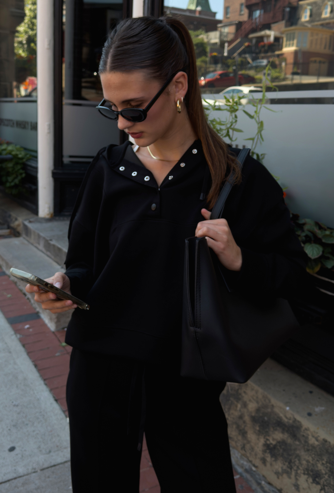 A woman wearing a black long-sleeve hoodie, black wide-leg pants, and black sunglasses, carrying a black tote bag on a city sidewalk.