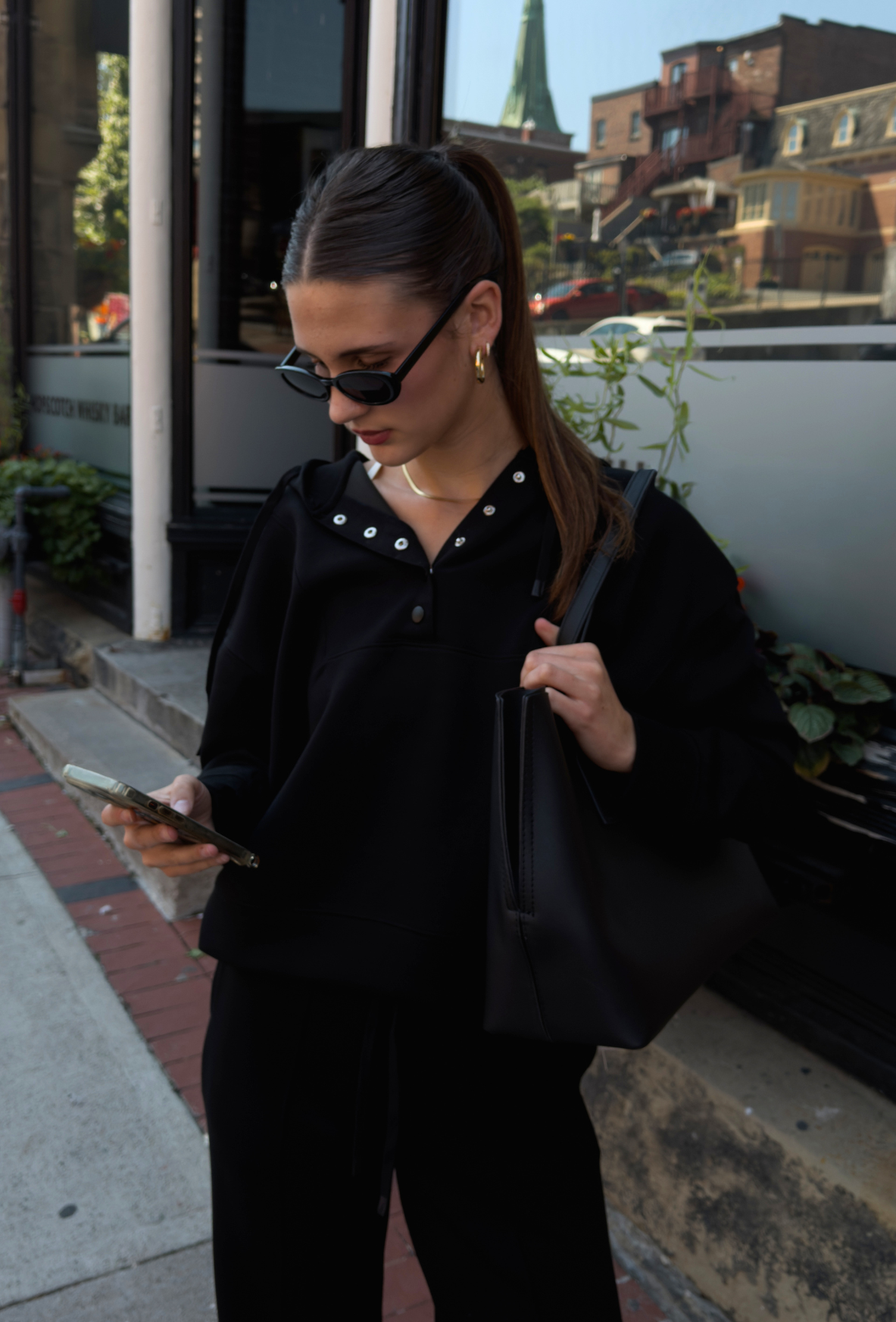A woman wearing a black long-sleeve hoodie, black wide-leg pants, and black sunglasses, carrying a black tote bag on a city sidewalk.