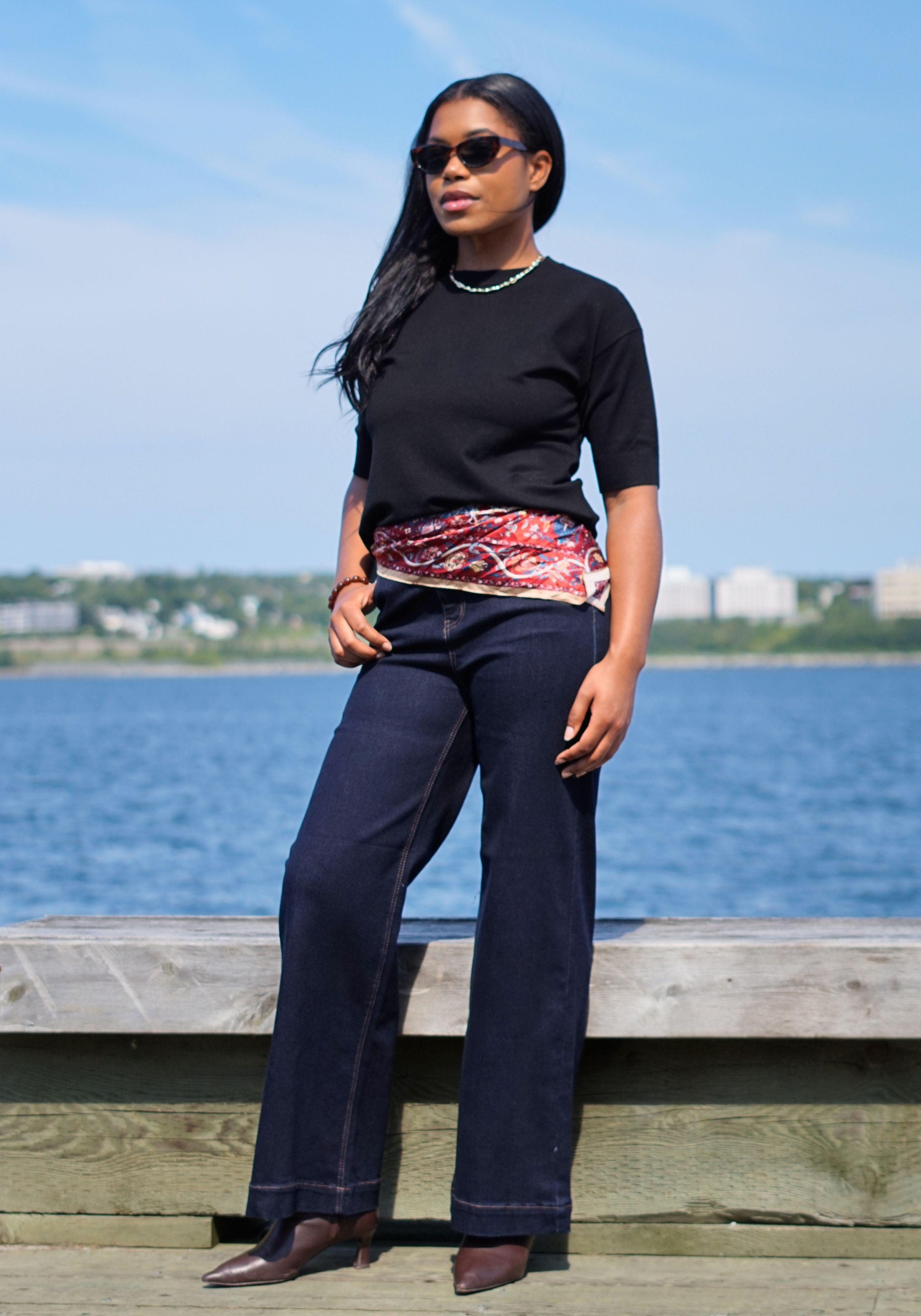 A woman wearing a black short-sleeve top, dark wide-leg jeans, sunglasses, and brown pointed shoes. A patterned red and pink sash is tied at her waist.