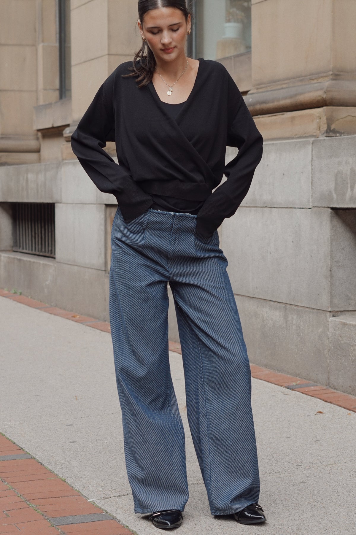 A woman wearing a black long-sleeve wrap-style top and wide-leg blue jeans, with her hands in her pockets.