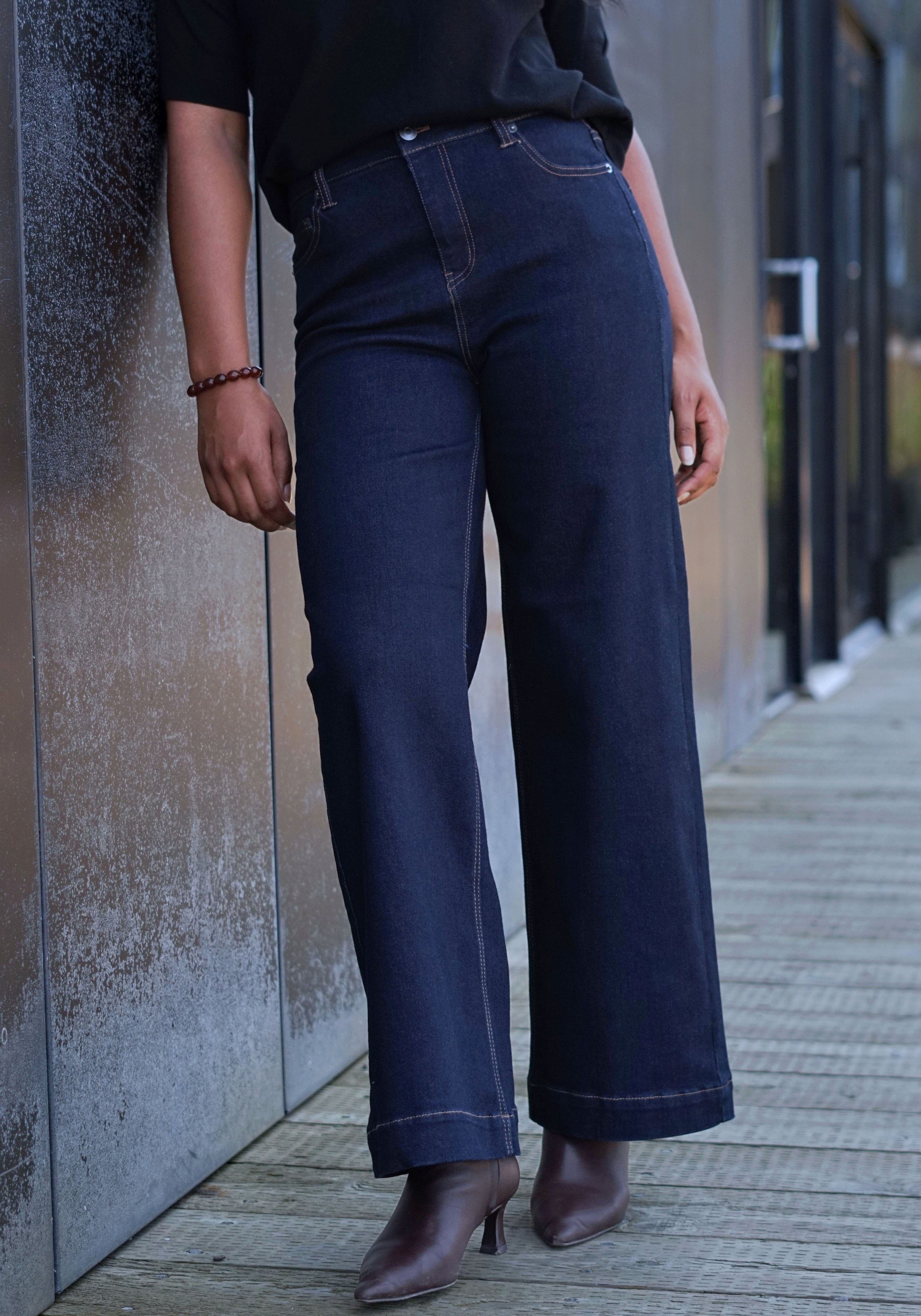 A person wearing dark blue wide-leg denim jeans, a black top, and brown ankle boots, standing outdoors next to a textured gray wall.