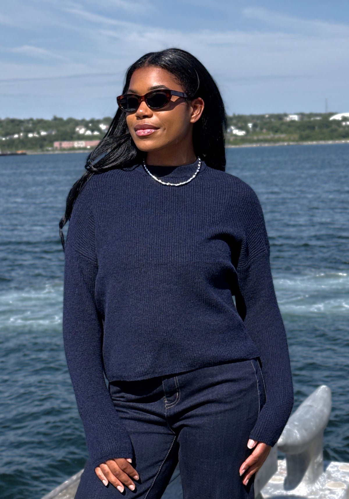 A woman wearing a dark navy blue ribbed knit mock neck sweater and matching navy pants stands outdoors by the water, wearing black sunglasses and a silver beaded necklace.