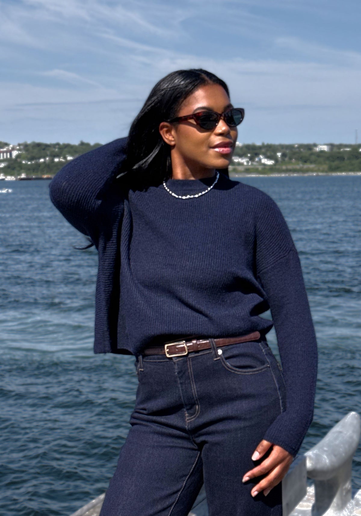 A woman wearing a dark navy ribbed knit long-sleeve sweater and dark blue jeans stands outdoors by the water, wearing sunglasses and a beaded necklace.
