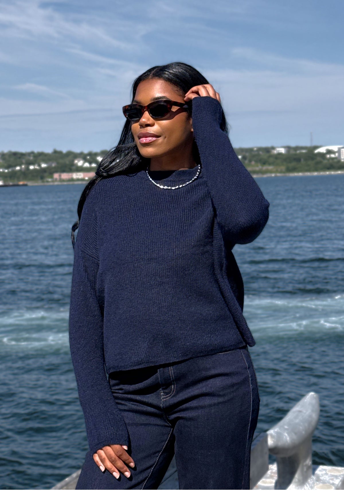 A woman wearing a dark navy blue ribbed knit sweater and dark wash denim pants stands outdoors by the water, wearing black sunglasses and a beaded necklace.