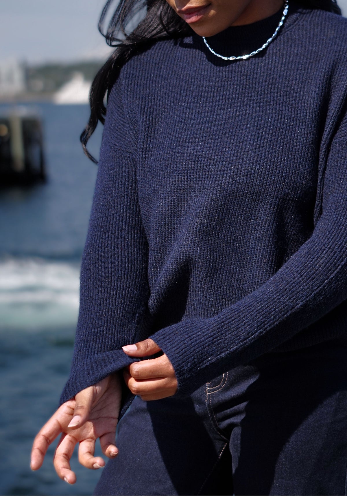 A close-up of a person wearing a dark navy ribbed knit sweater outdoors near the water.