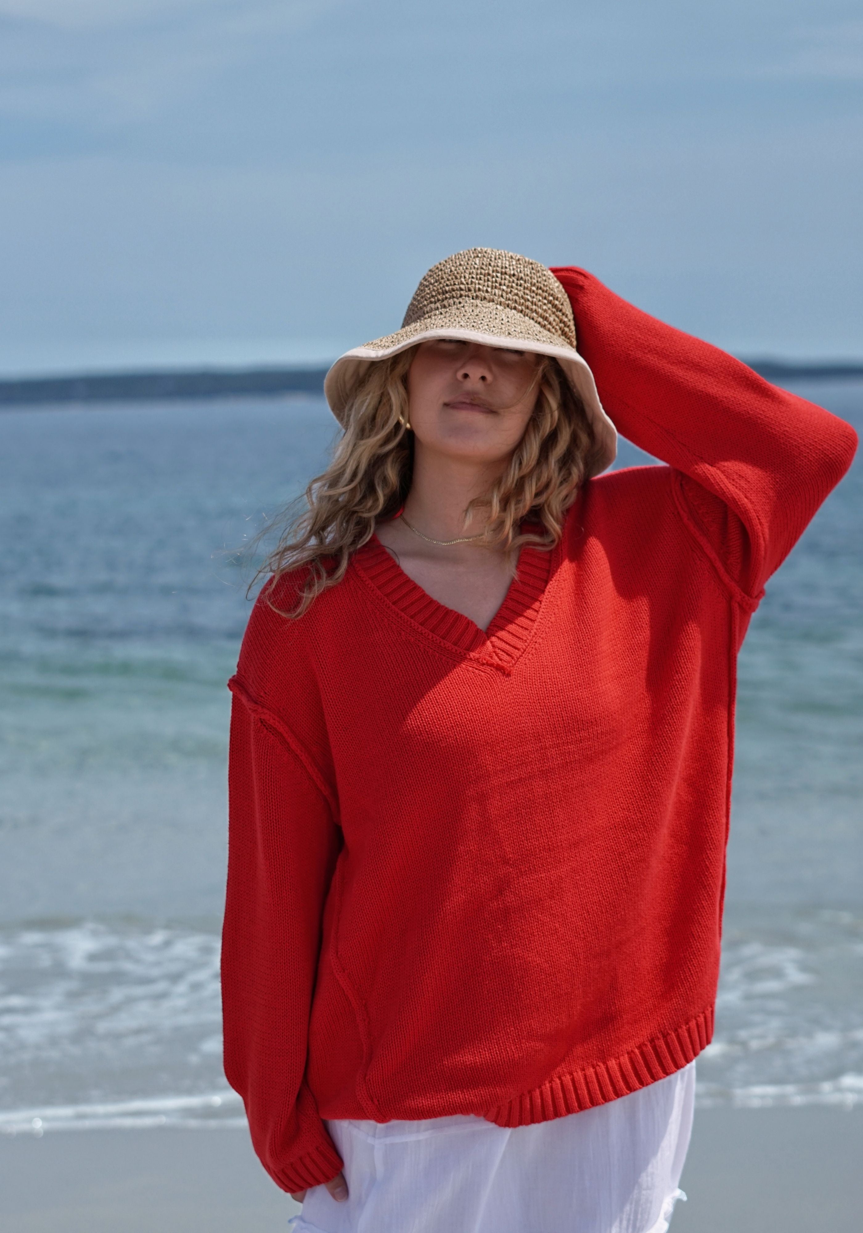 A woman standing on a beach wearing a bright red knit V-neck sweater, a straw sun hat, and white bottoms, with the ocean and sky in the background.