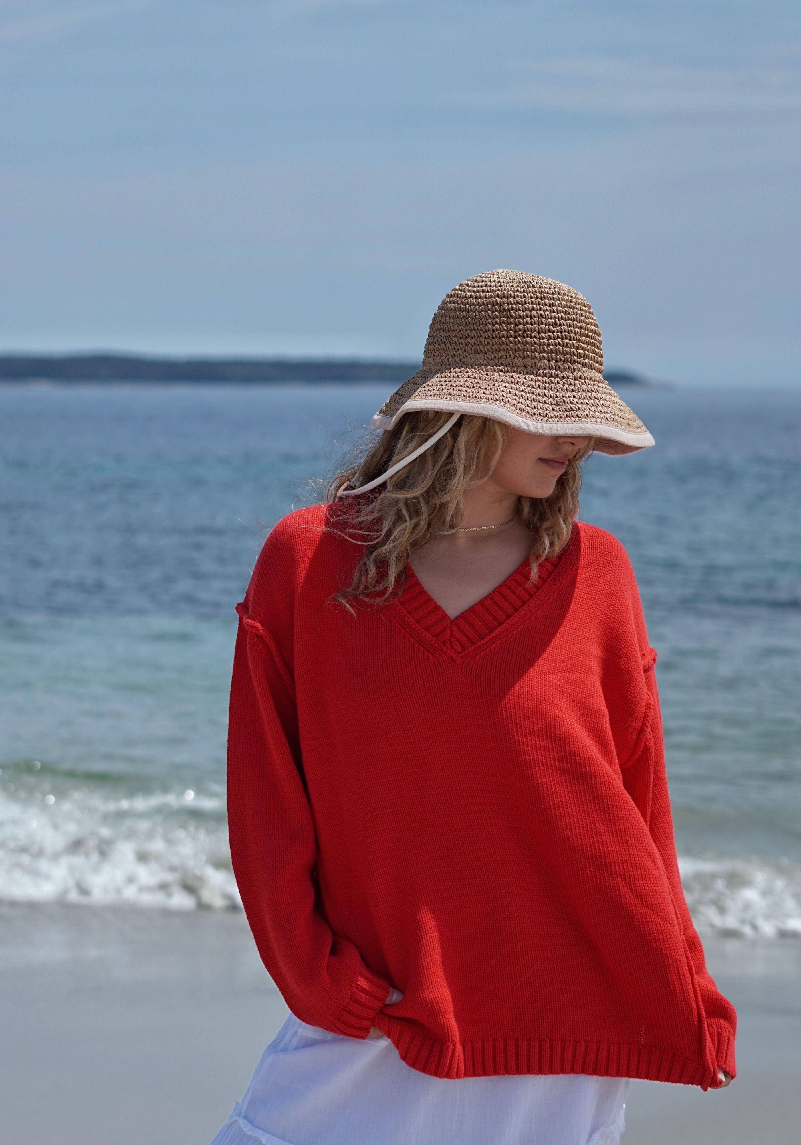 A woman standing on a beach wearing a bright red knit V-neck sweater, a beige straw sun hat, and white bottoms.