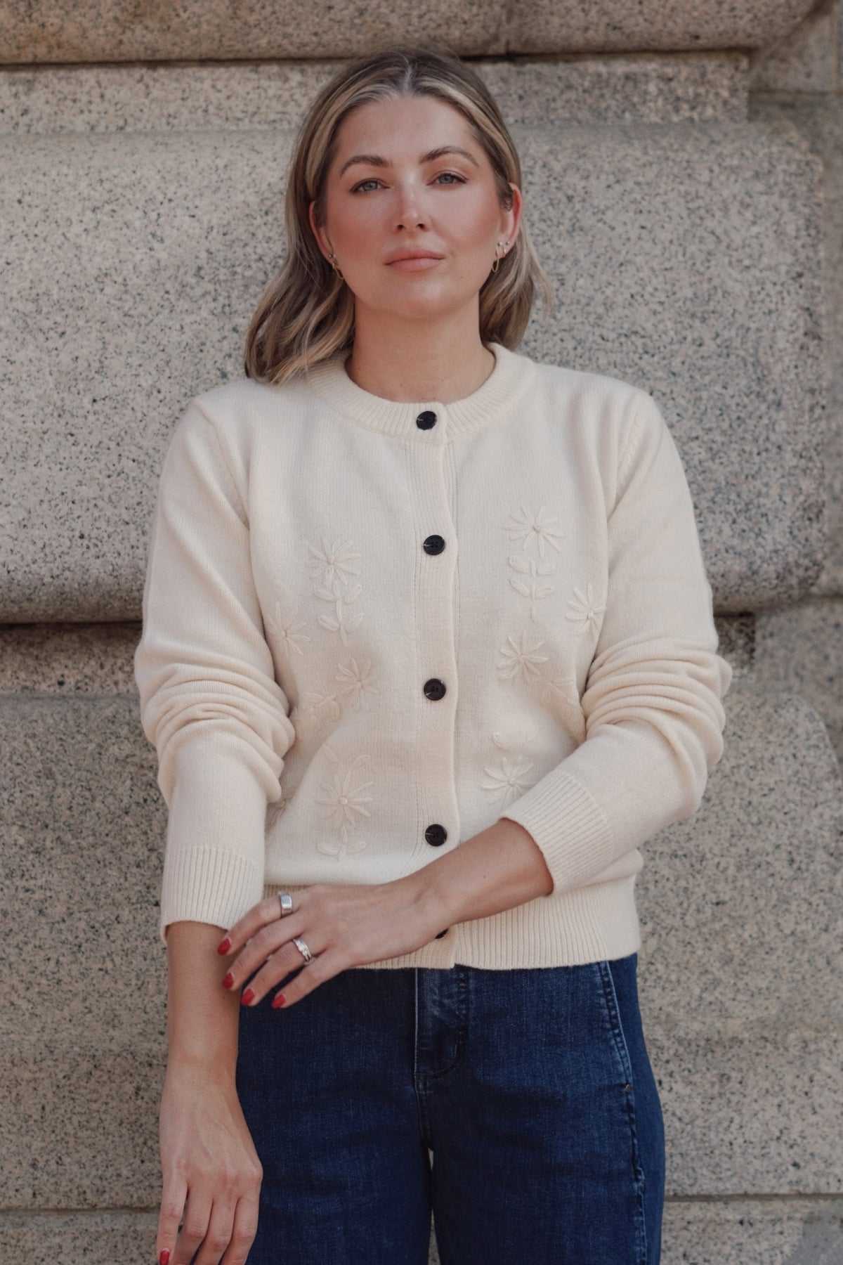 A woman wearing a cream-colored knit cardigan with black buttons and subtle floral embroidery, paired with dark blue jeans