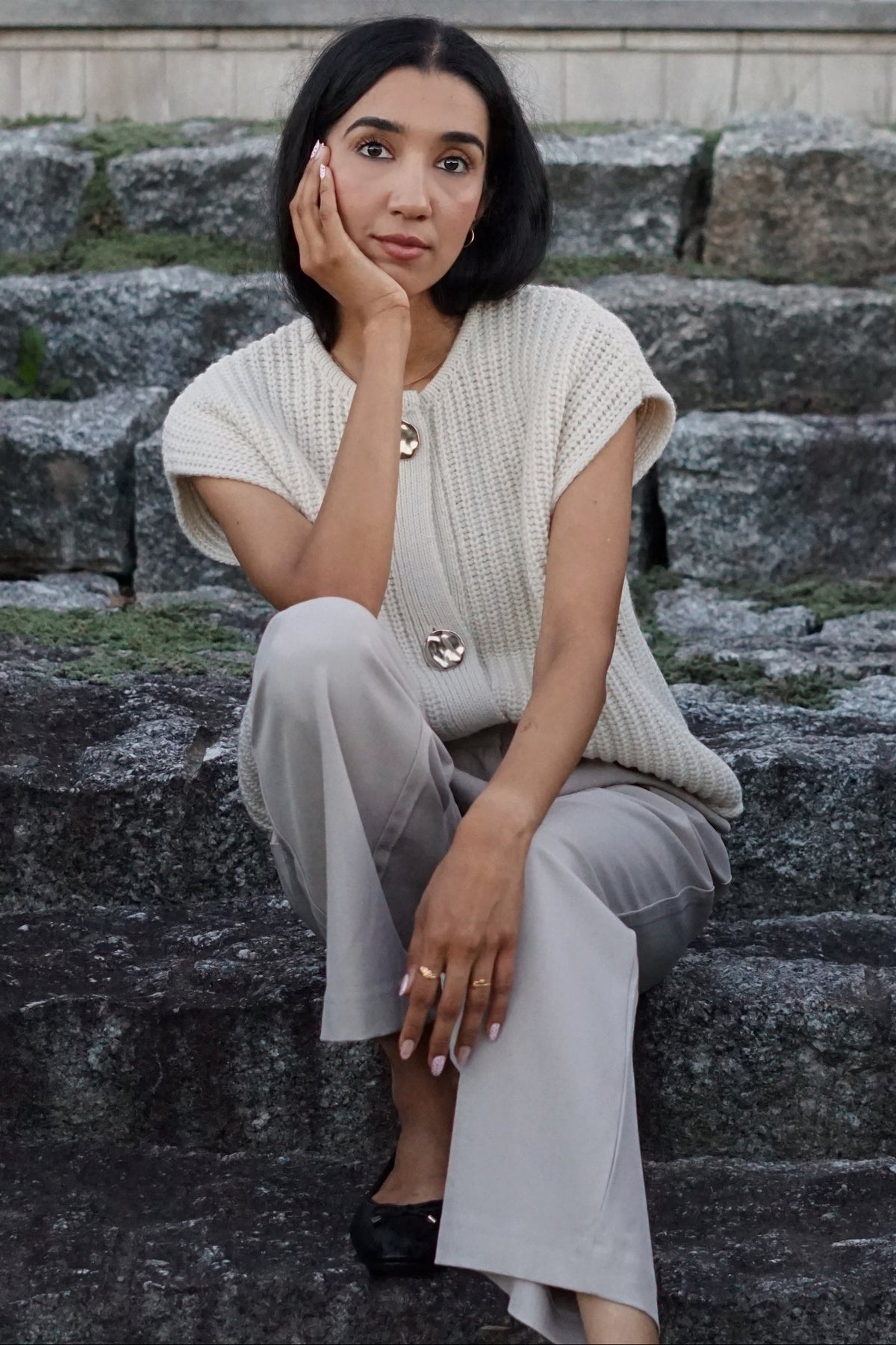 A woman sits on stone steps wearing a cream ribbed knit vest with decorative buttons and light gray wide-leg trousers.