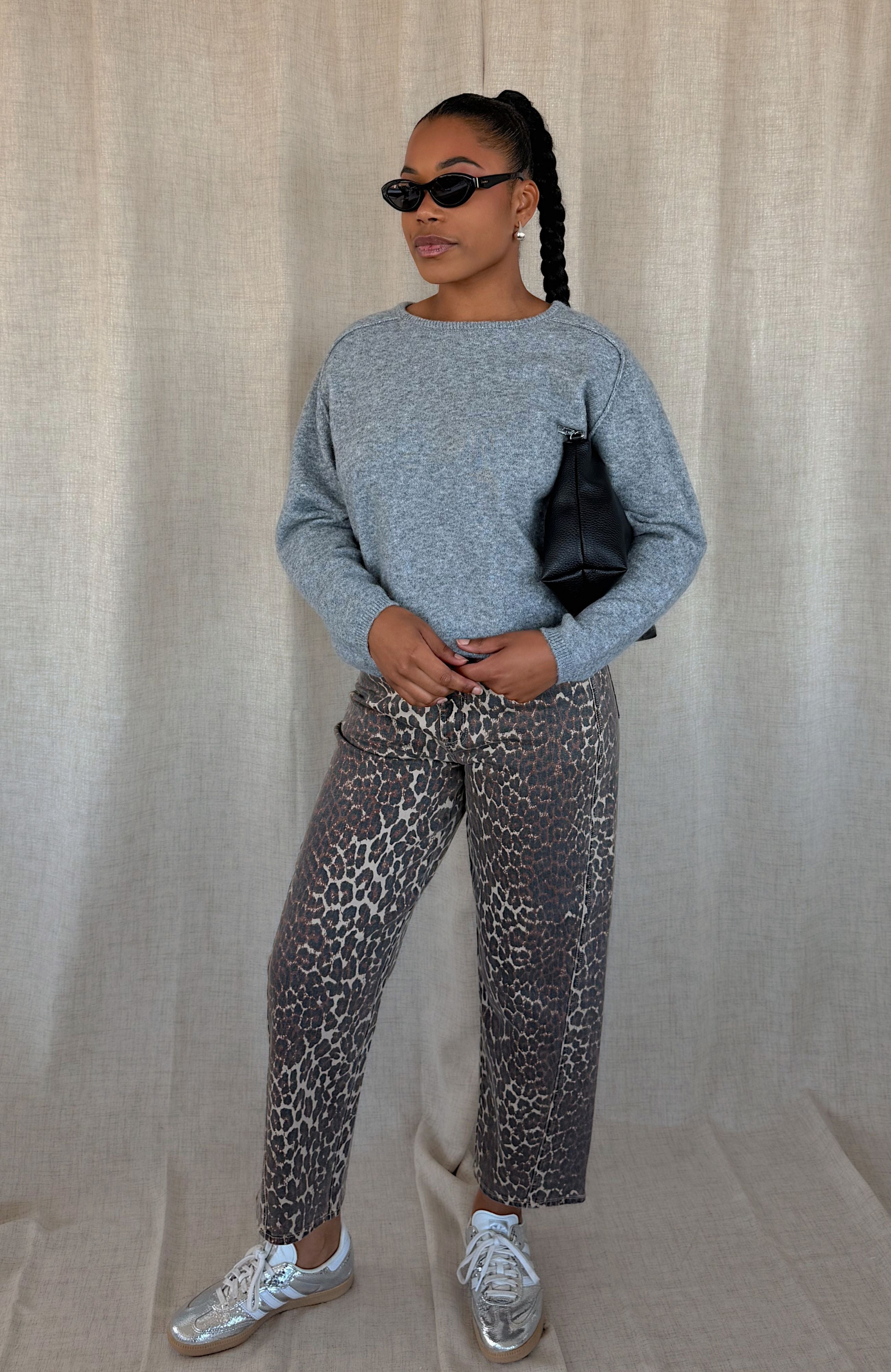 A woman modeling a light gray long-sleeve knit sweater and leopard-print pants, styled with black sunglasses, a black shoulder bag, and white sneakers, standing against a beige fabric backdrop.