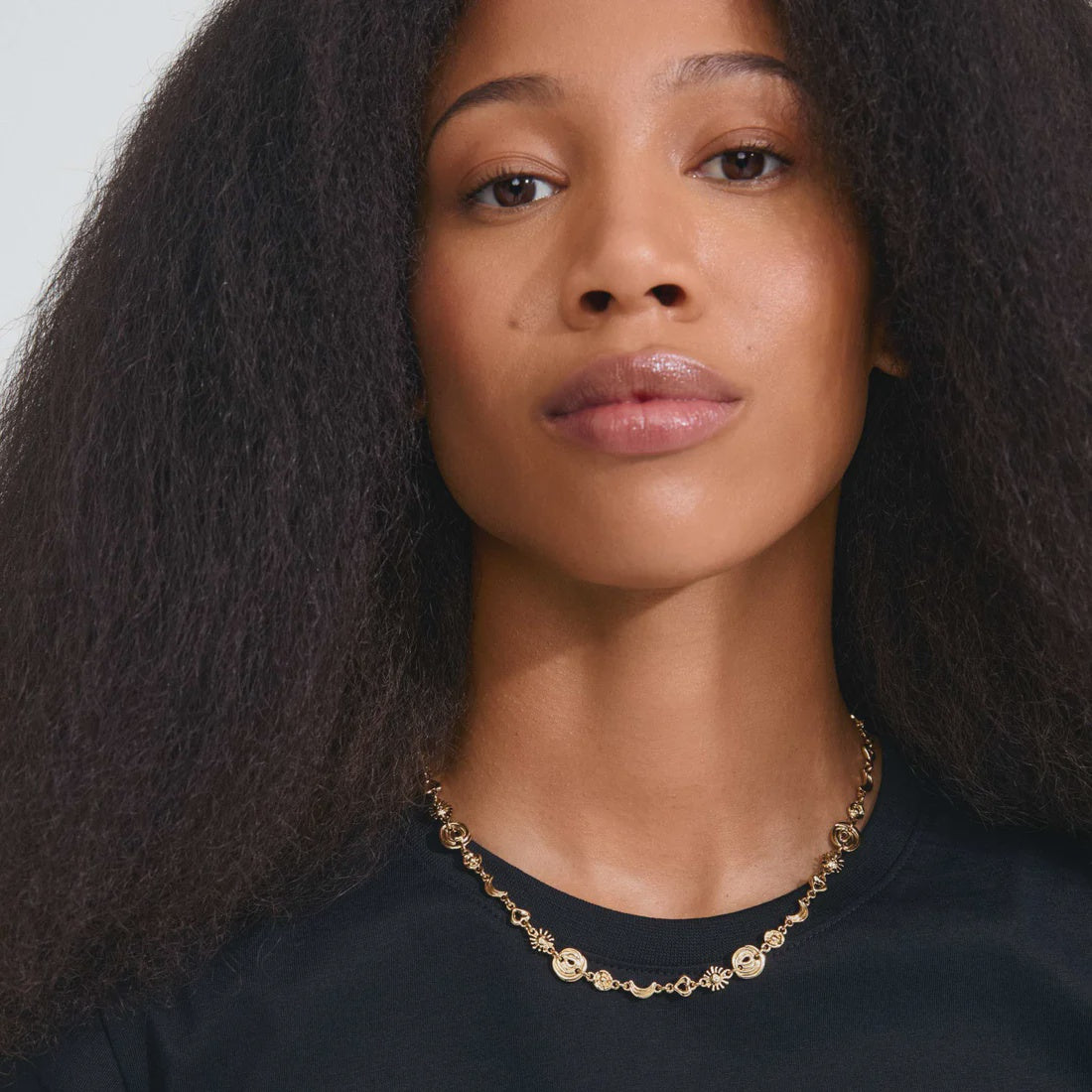 A close-up portrait of a woman with long, dark, curly hair wearing a delicate gold chain necklace with small circular charms, paired with a black top against a light gray background.