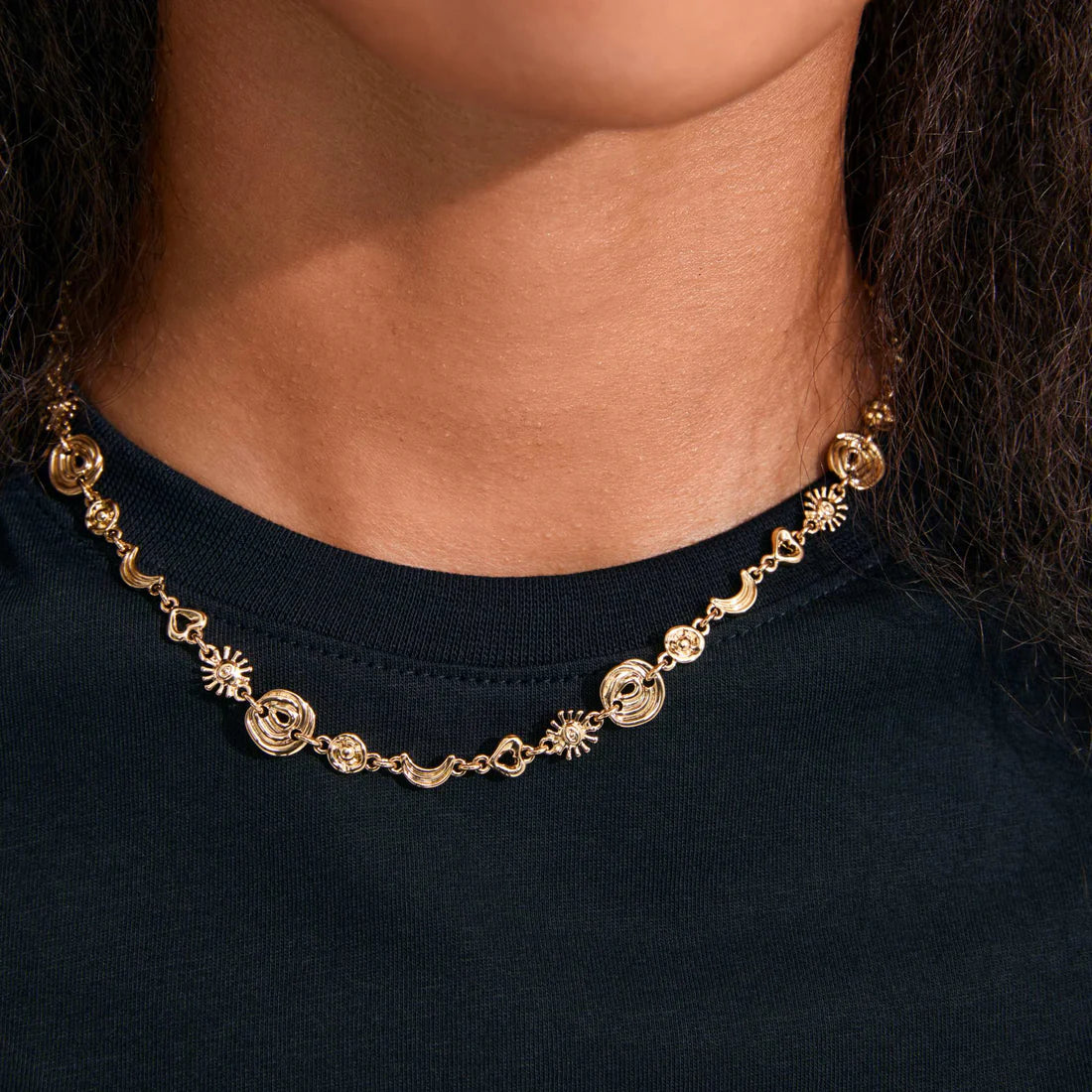 A close-up of a person wearing a delicate gold chain necklace with celestial charms, including suns, moons, and a central medallion, styled with a black crewneck shirt.