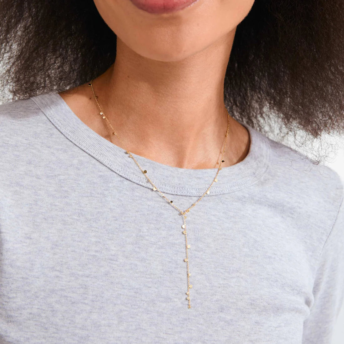 A close-up of a model wearing a delicate gold Y-necklace with tiny dangling charms over a light gray heathered t-shirt.