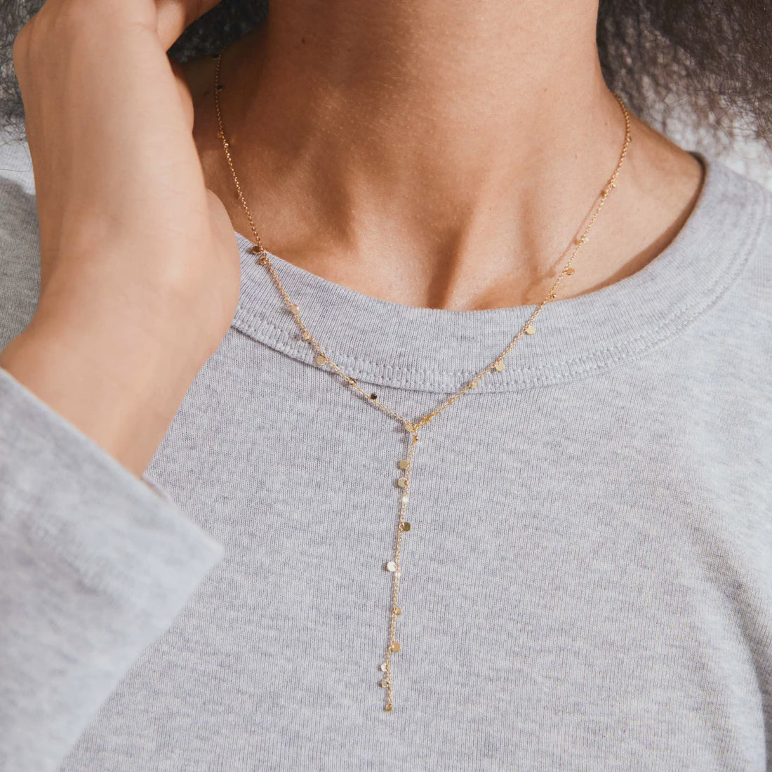 A close-up of a model wearing a delicate gold Y-necklace with small star-shaped charms over a light gray heathered t-shirt.
