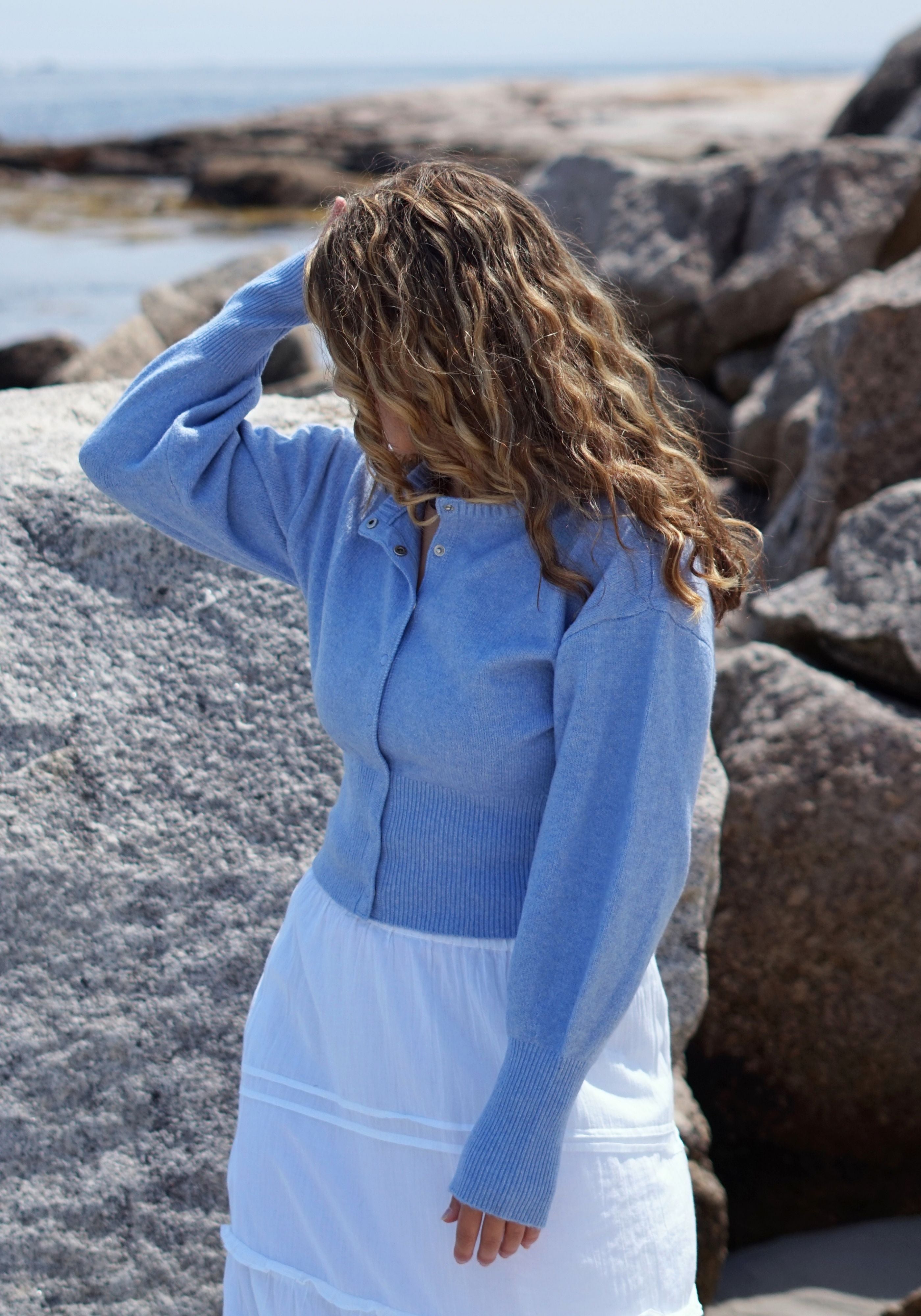 A woman standing outdoors on a rocky beach wearing a light blue knit cardigan and a white skirt