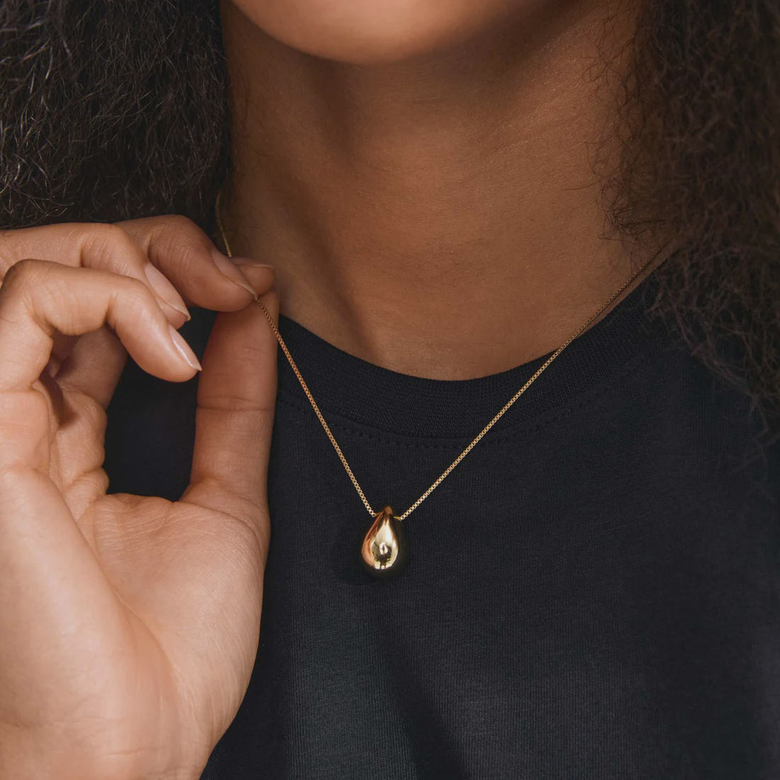 A close-up of a person wearing a delicate gold chain necklace with a small teardrop-shaped gold pendant, styled with a black crew-neck t-shirt.