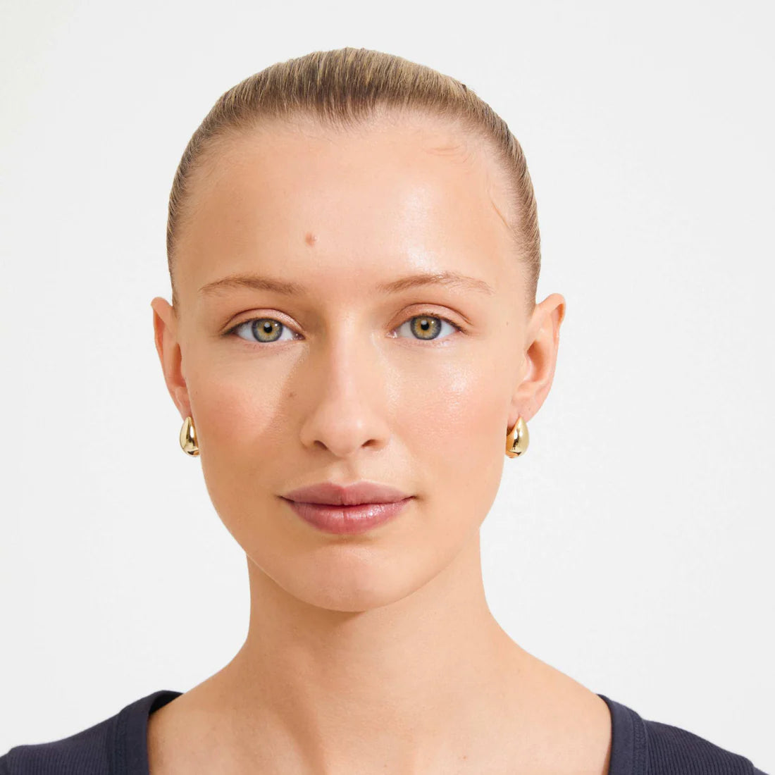 A close-up portrait of a woman with her hair pulled back, wearing small gold hoop earrings and a dark navy top against a plain light gray background.