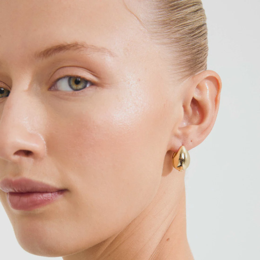 A close-up of a model's ear wearing a small, polished gold teardrop-shaped stud earring against a clean white background.