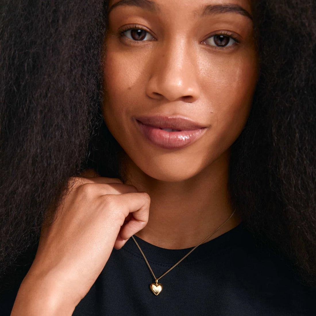 A close-up portrait of a woman with dark hair wearing a delicate gold heart pendant necklace on a dark navy top, set against a dark background.