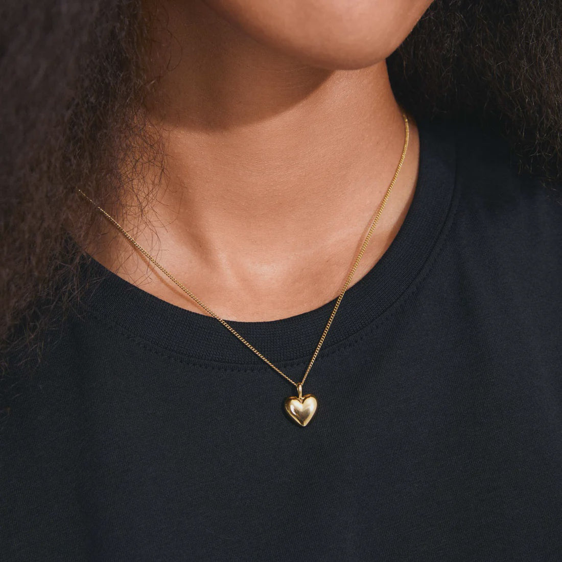 A close-up of a person wearing a delicate gold heart pendant necklace on a fine chain, paired with a black crewneck shirt.