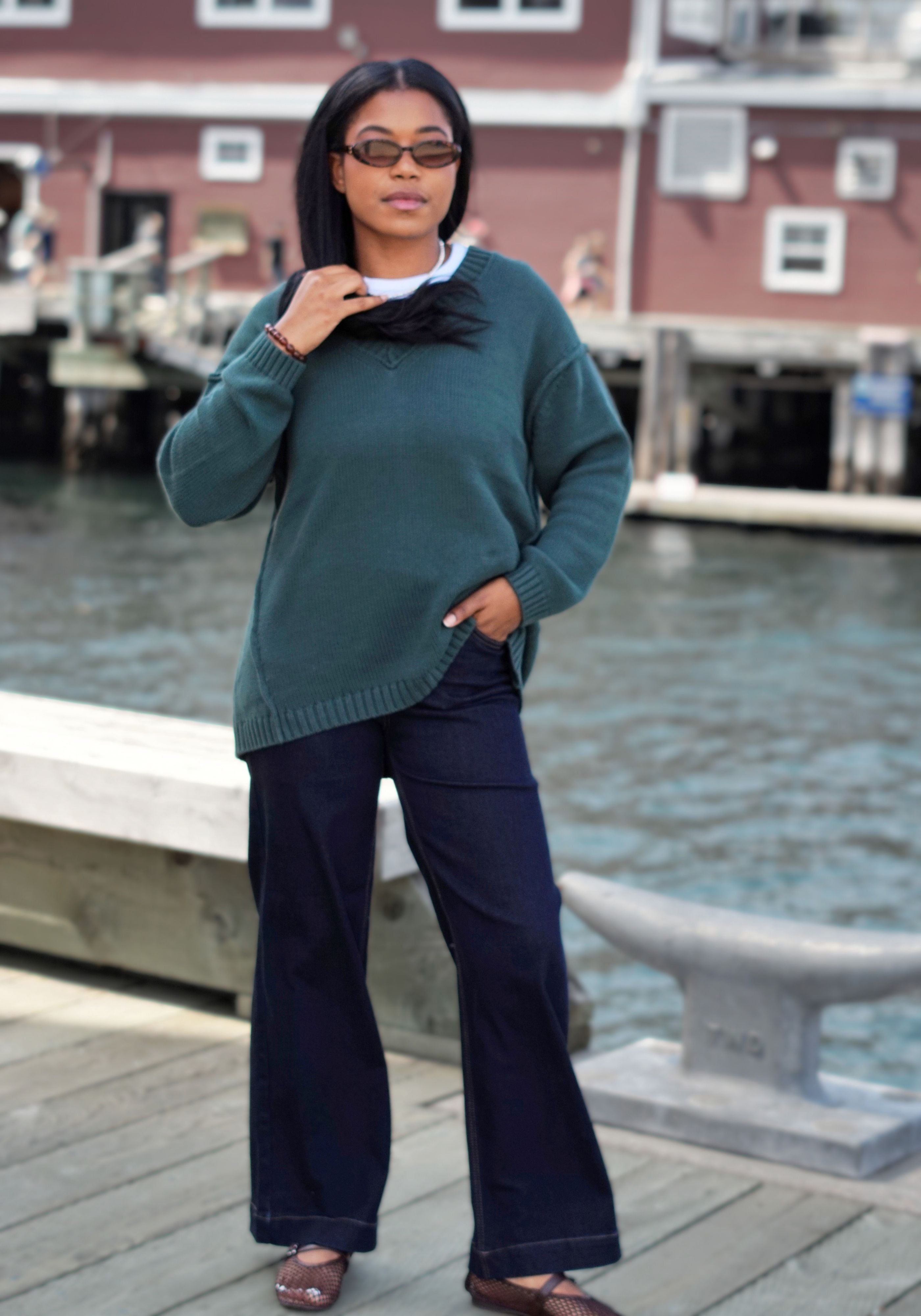 A woman stands on a wooden dock wearing a dark teal knit sweater, navy wide-leg pants, and brown shoes.