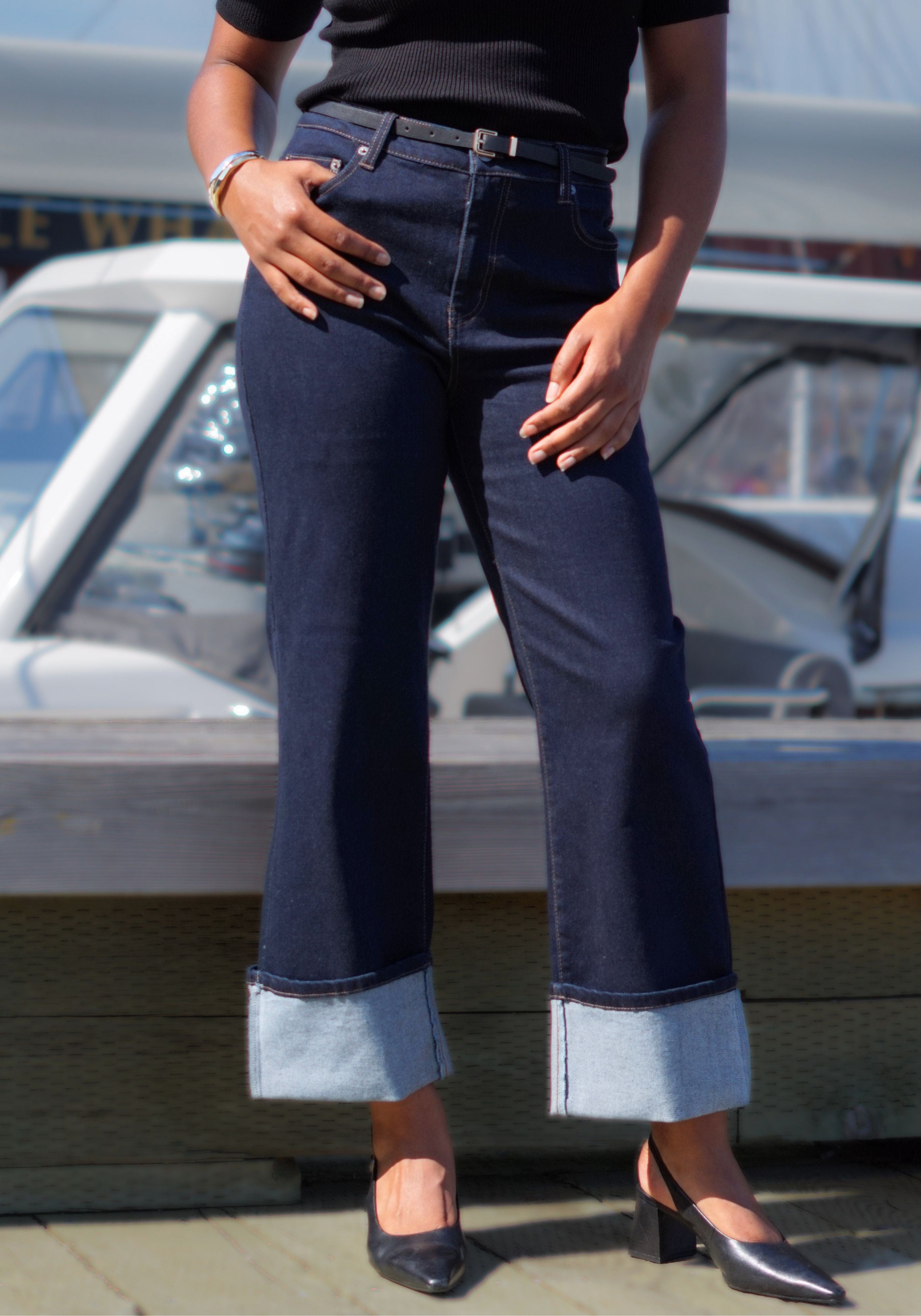 A person wearing dark blue cropped jeans with rolled cuffs, paired with a black top and black heels, standing outdoors.