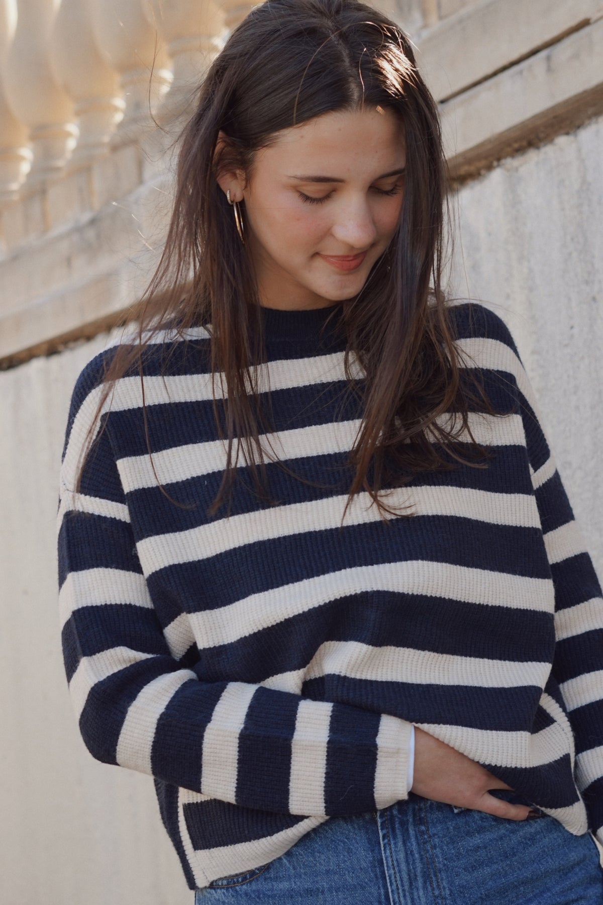 A woman wearing a navy blue and off-white horizontally striped knit sweater, paired with blue jeans, standing against a light-colored stone wall.