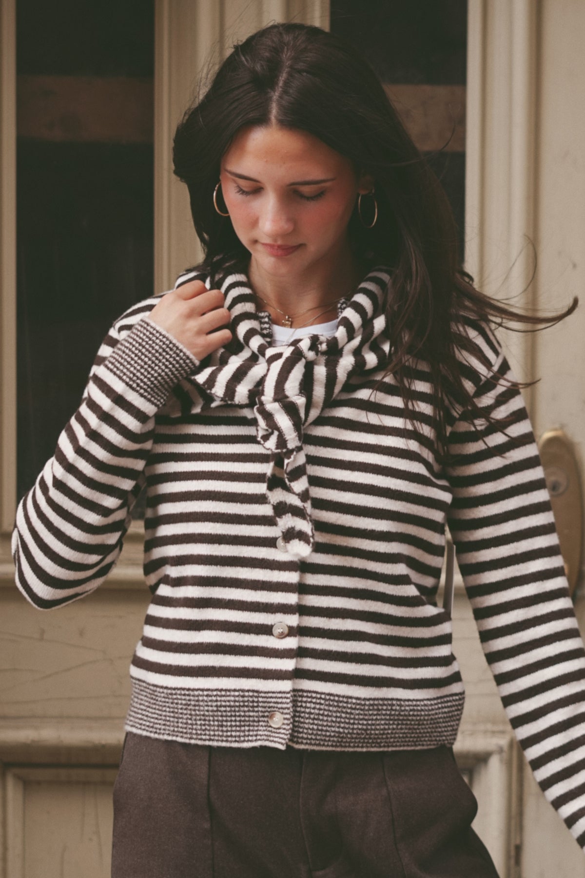 A woman wearing a black and white horizontally striped knit cardigan with a matching knotted scarf, paired with dark brown pants, standing in front of a rustic wooden door.