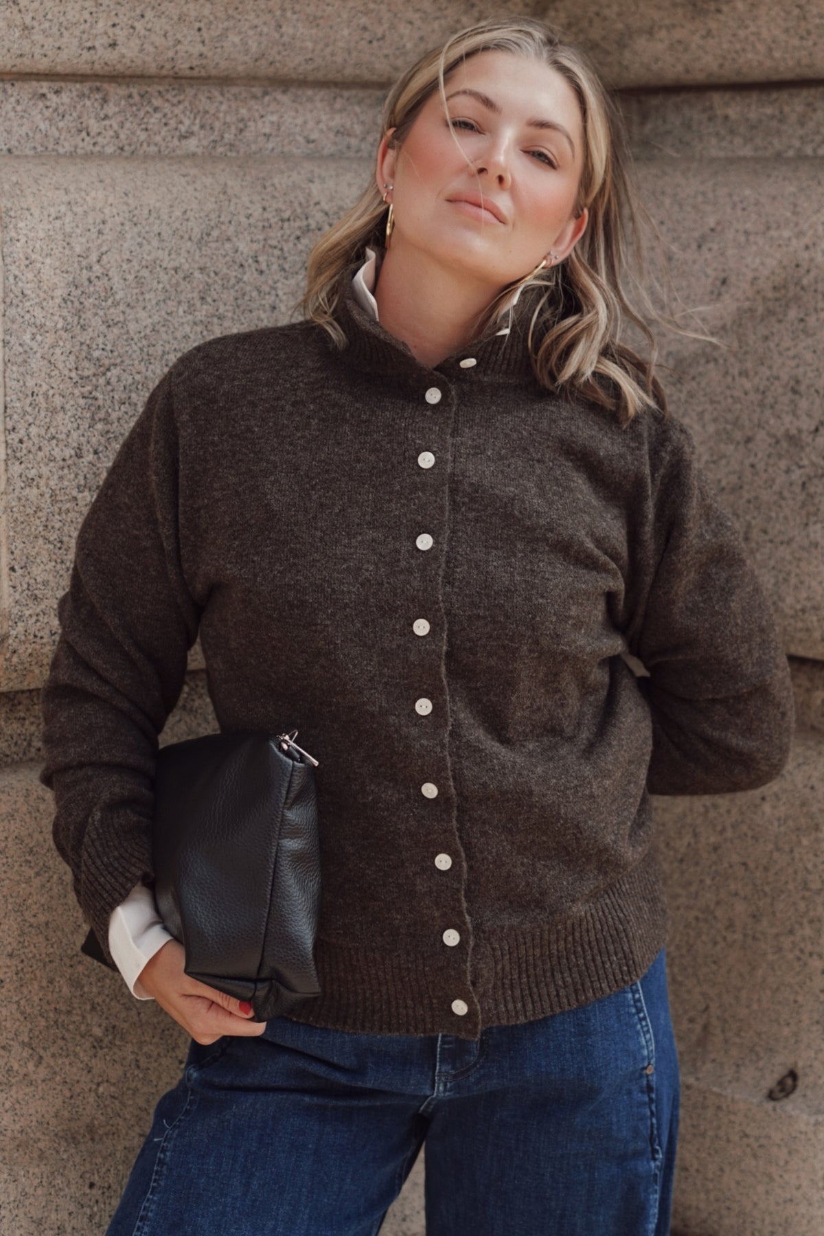 A woman wearing a dark brown button-up knit cardigan sweater and blue jeans, holding a black clutch bag, standing against a textured stone wall.