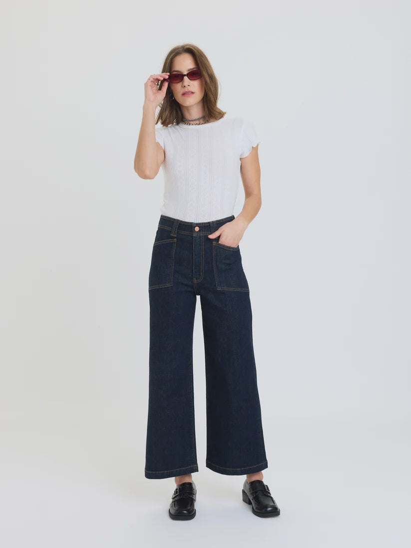A woman modeling a white short-sleeve ribbed top and dark wide-leg denim jeans with black shoes, standing against a plain white studio background.