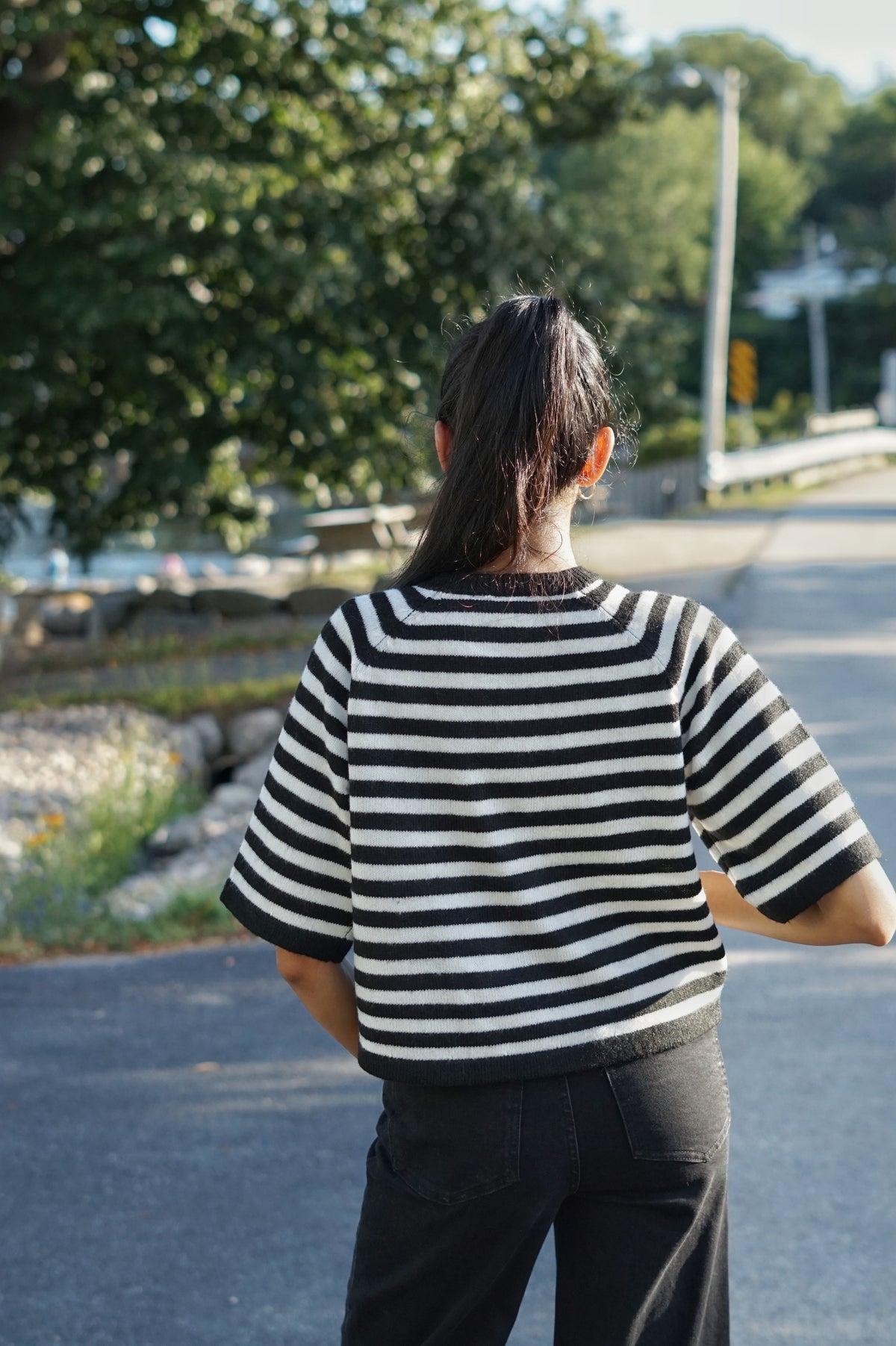 A person viewed from behind wearing a black and white horizontally striped short-sleeve knit top and dark pants, standing on an outdoor paved path with trees in the background.