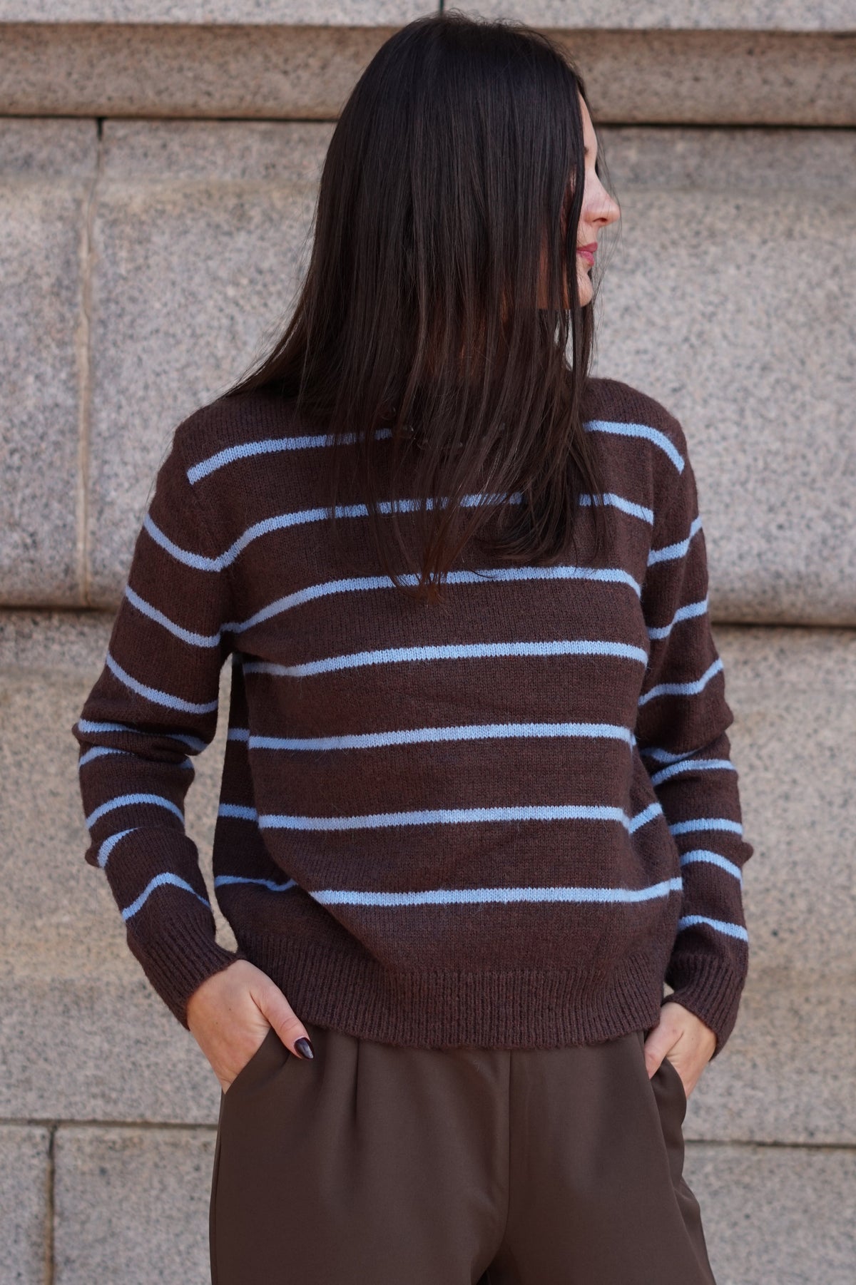 A woman wearing a brown knit sweater with light blue horizontal stripes and brown trousers, standing against a stone wall.