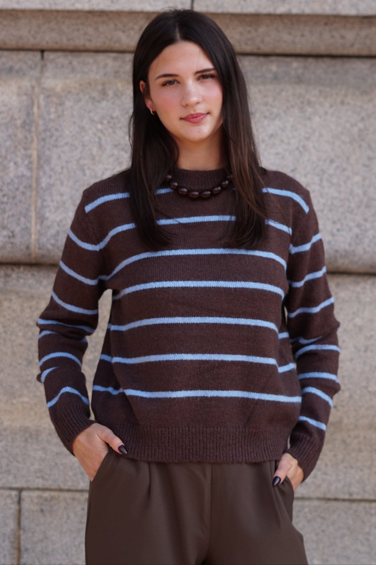 A woman wearing a brown knit sweater with light blue horizontal stripes and brown pants.