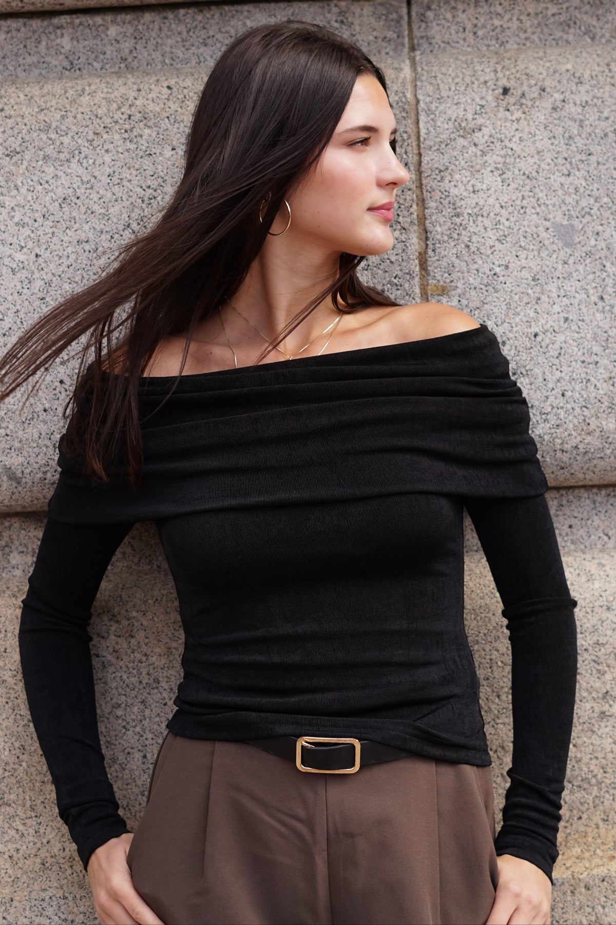 A woman wearing a black off-the-shoulder long-sleeve top and brown trousers, standing against a gray stone wall.