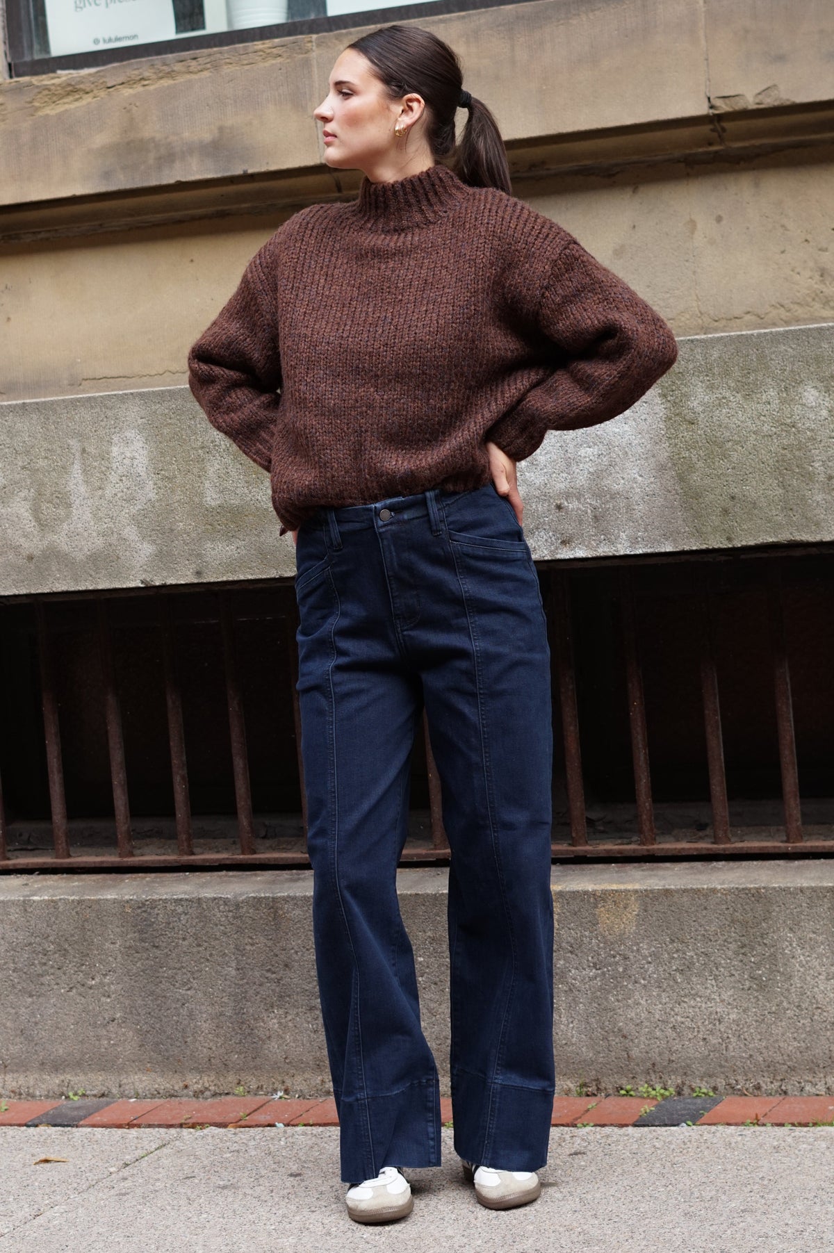 A woman wearing a brown ribbed mock-neck sweater and dark blue wide-leg jeans, standing outdoors against a stone building.