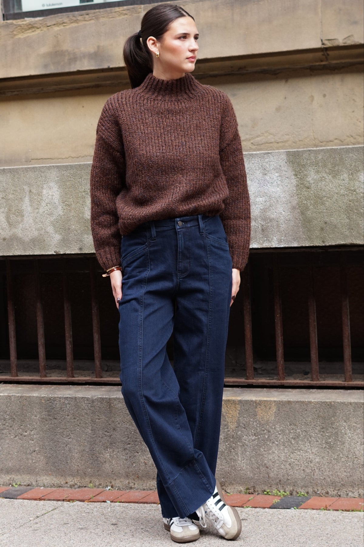 A woman wearing a brown ribbed knit mock neck sweater, dark blue wide-leg jeans, and silver sneakers, standing outdoors against a concrete wall.