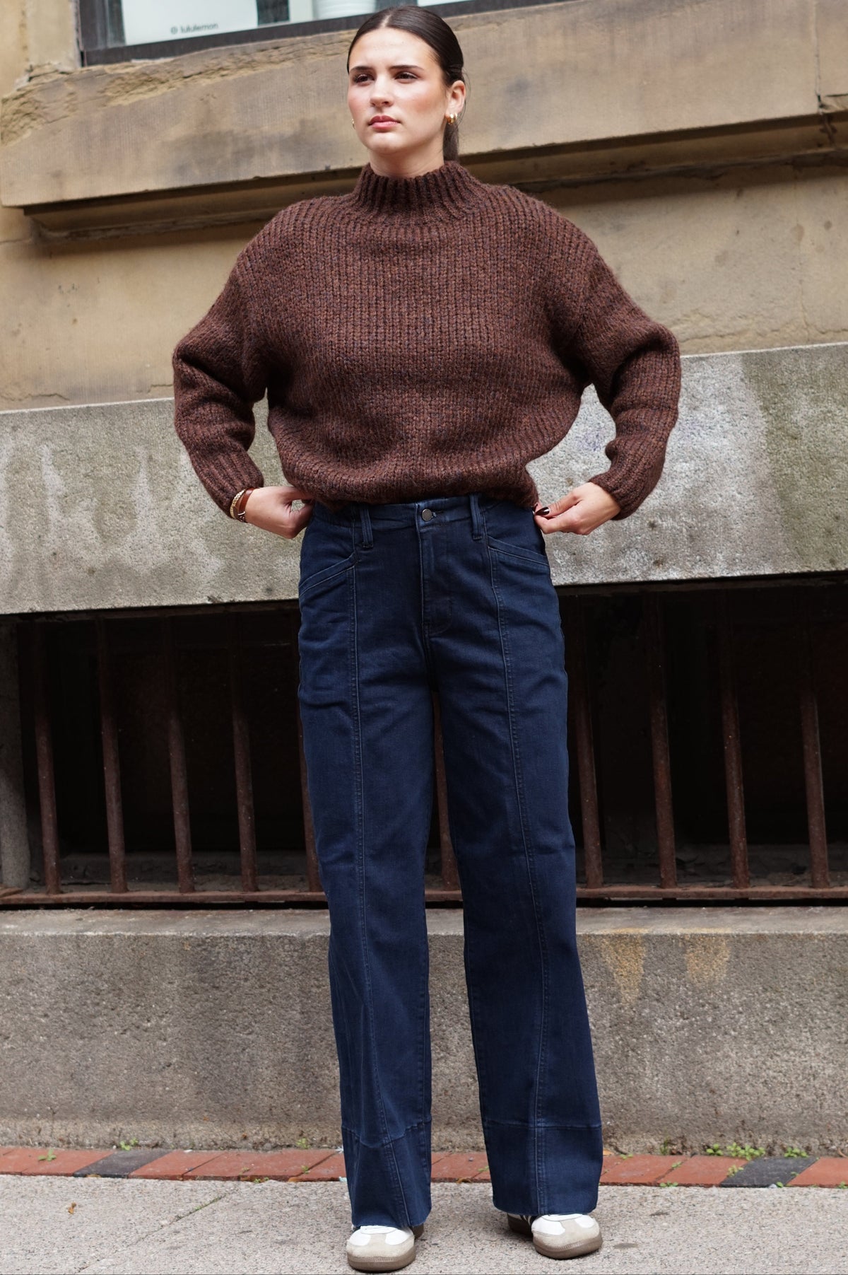 A woman stands outdoors in front of a stone building, wearing a brown ribbed knit mock-neck sweater and dark blue wide-leg jeans with white sneakers.