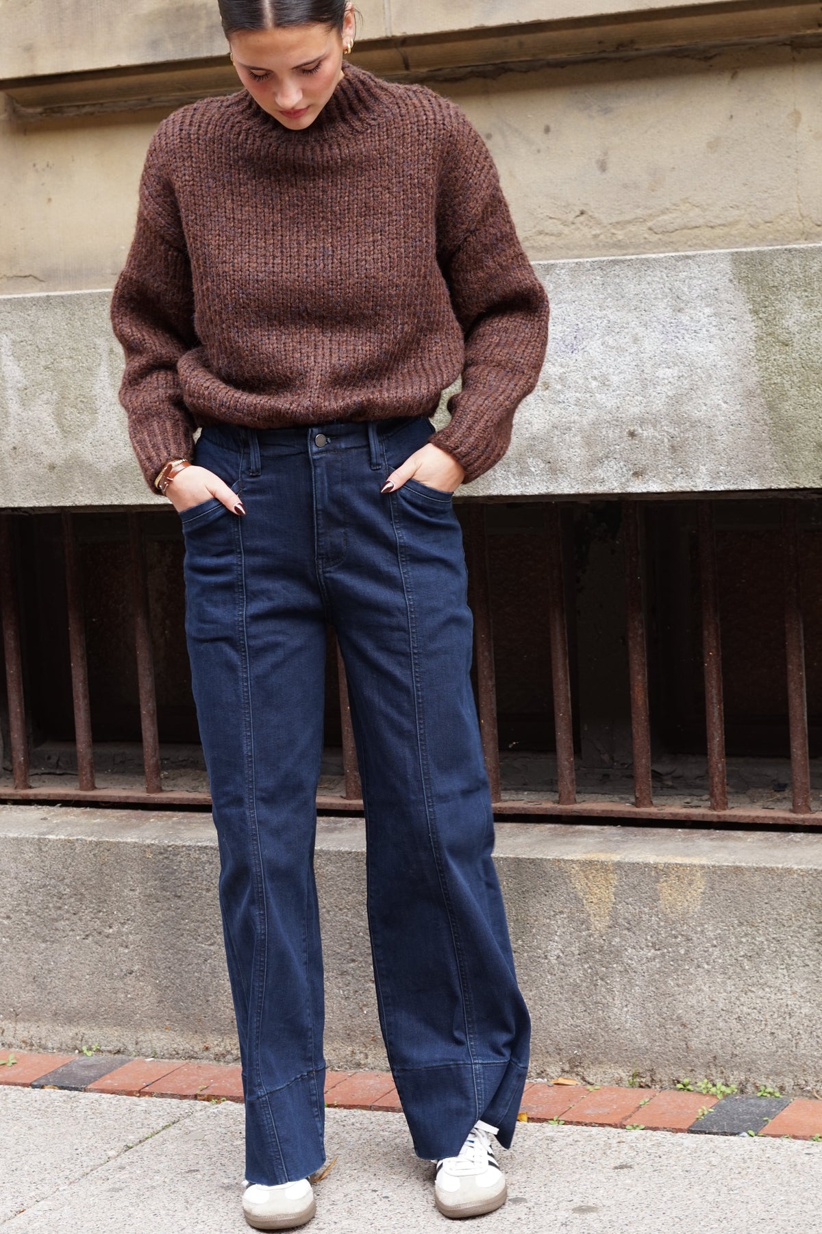 A woman wearing a brown ribbed knit sweater and dark blue wide-leg jeans, standing outdoors against a concrete wall.