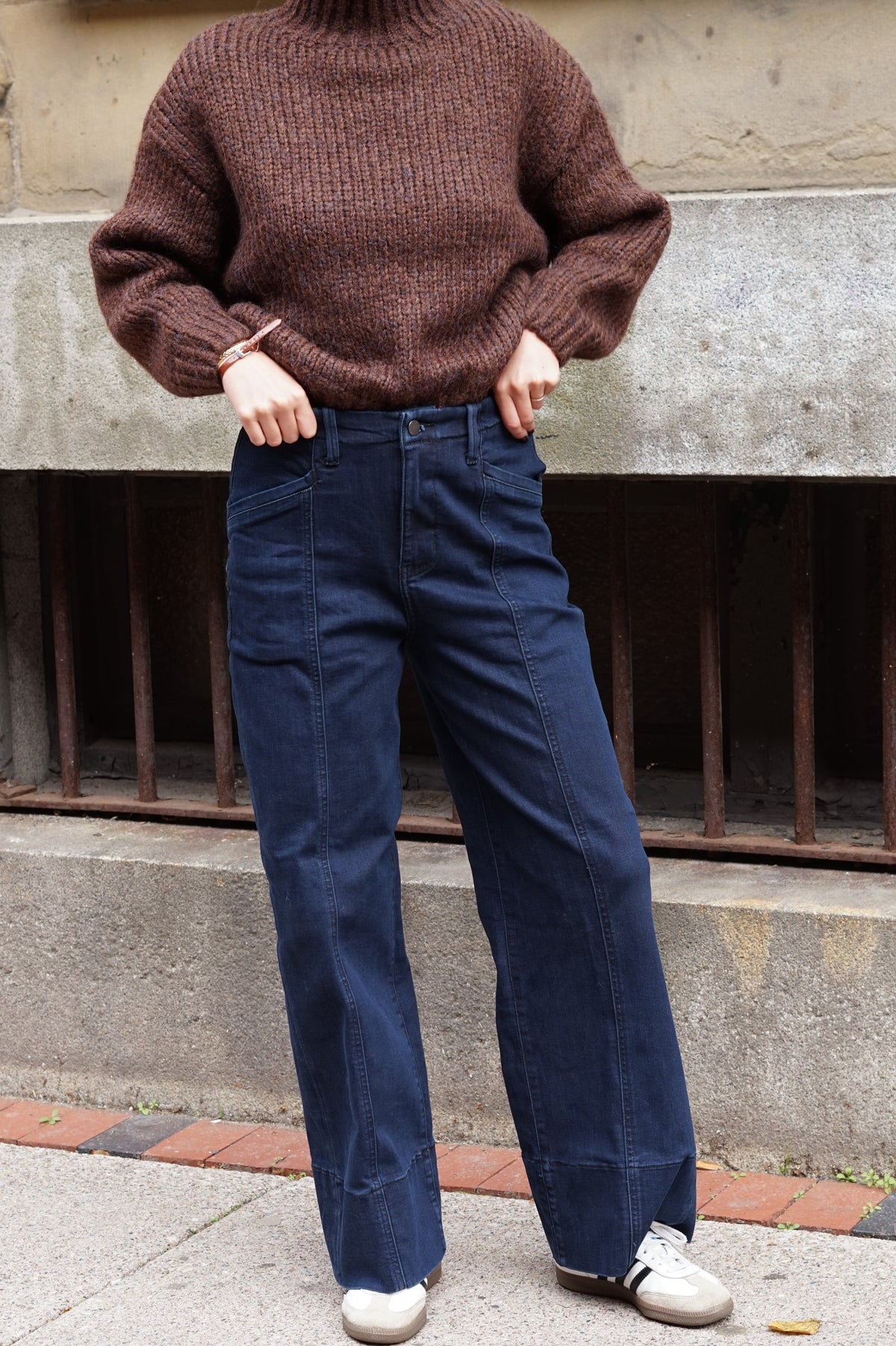 A person wearing a brown ribbed knit turtleneck sweater and dark blue wide-leg jeans, standing outdoors in front of a concrete wall and metal gate.