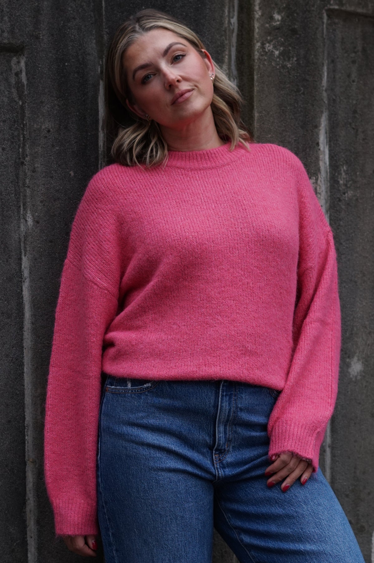 A woman wearing a bright pink knit crewneck sweater and blue jeans, leaning against a dark textured concrete wall.